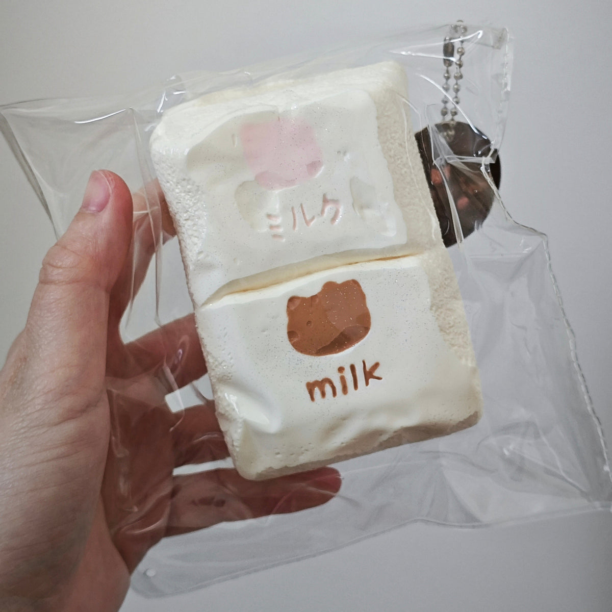 A hand holding a clear plastic bag containing a small novelty item shaped like a cream-colored marshmallow or sponge cake with pink and brown printed details and the word 'milk' on it.