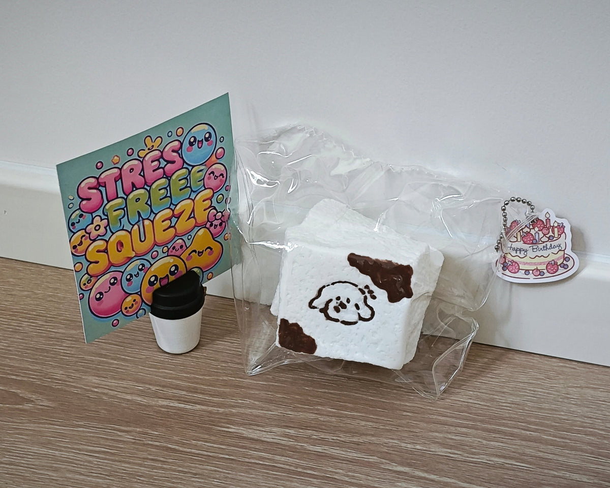 A cute white squishy toy shaped like a slice of cake with brown chocolate drizzle and a cartoon dog face, shown in clear plastic packaging next to a small coffee cup and a colorful 'Stress Free Squeeze' card on a wooden floor.