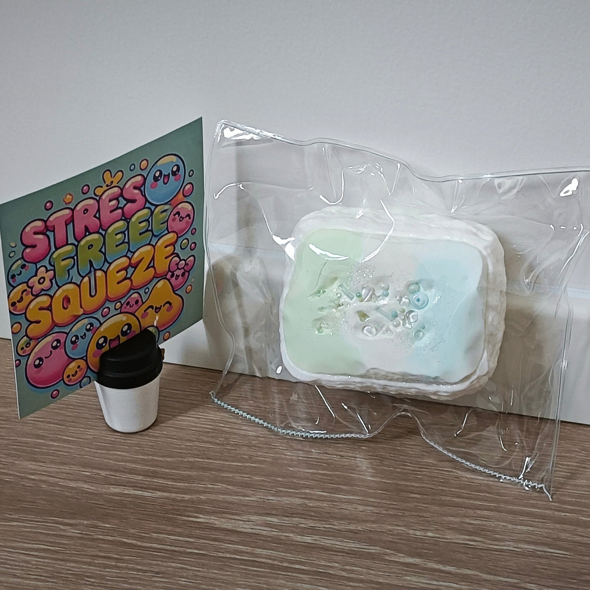 A clear plastic bag containing a pale mint and white rectangular item with embossed floral details, placed on a wood floor against a white wall. A colorful card reading 'STRESS FREE SQUEEZE' and a small black-and-white cup sit beside it.