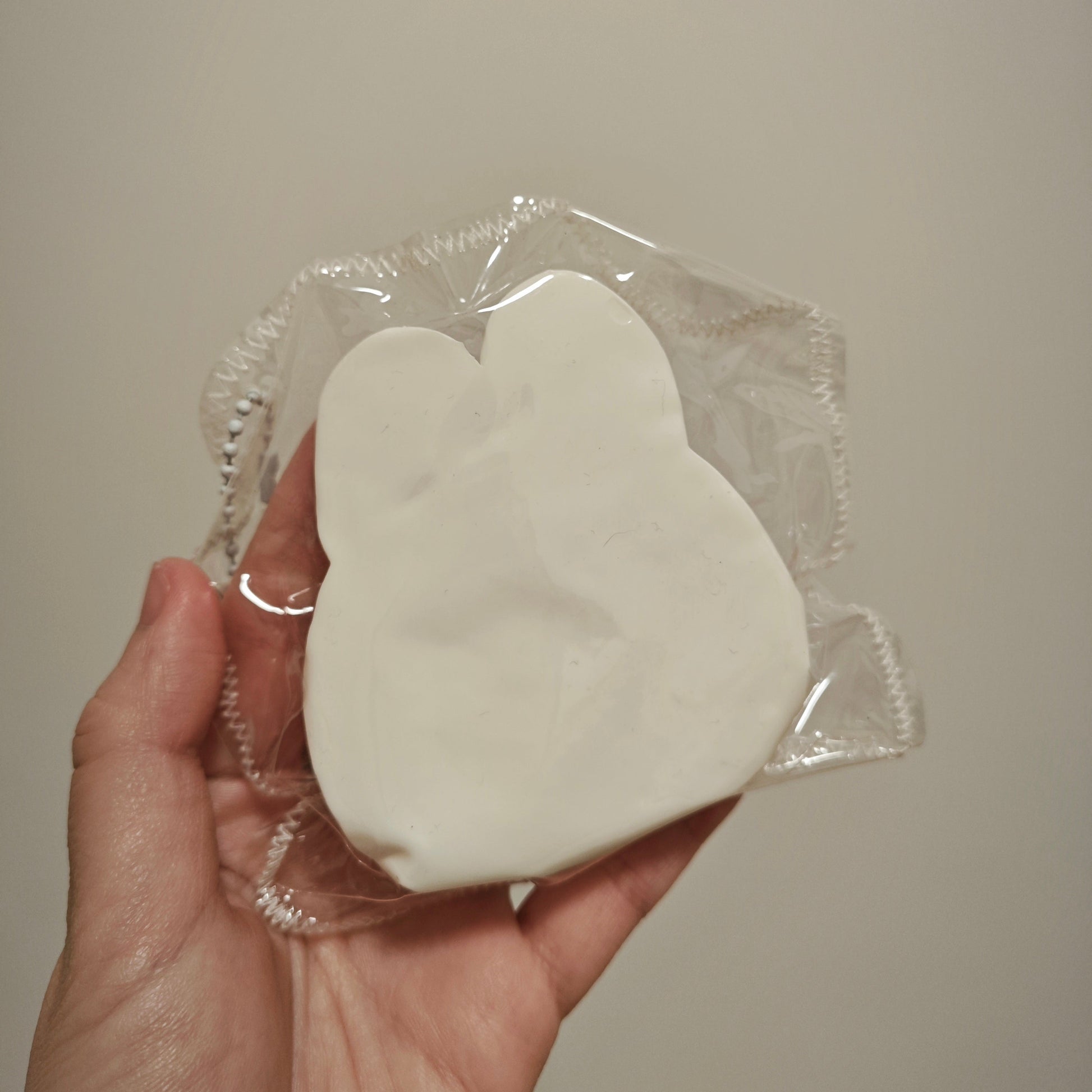 A hand holding a white, bunny-shaped squishy toy wrapped in clear plastic against a plain beige background.