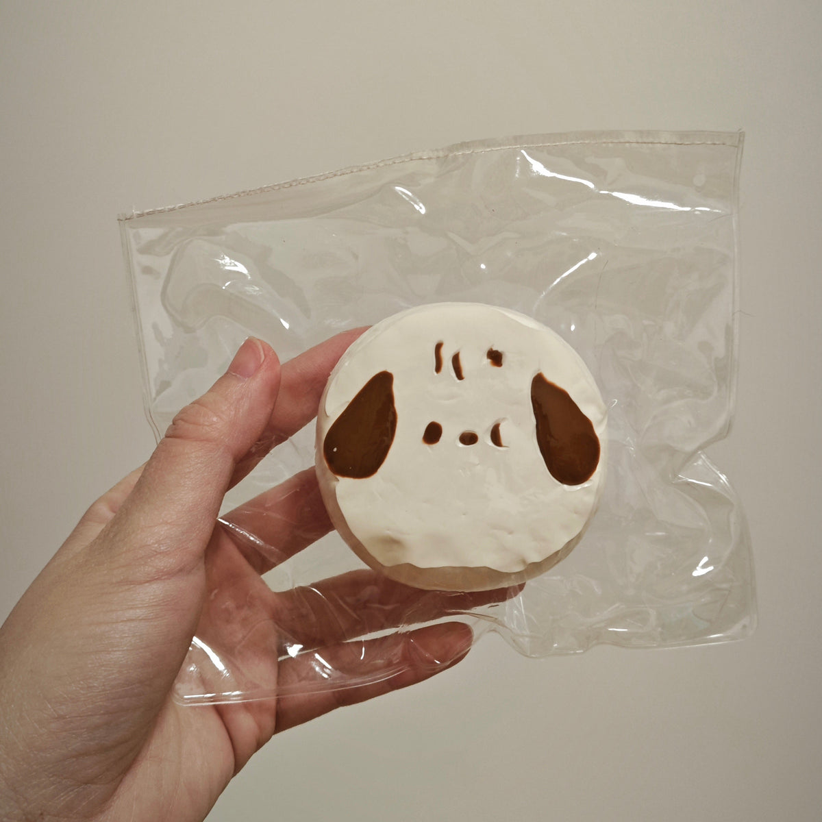 A hand holding a small round white item with brown ear-like shapes and simple facial markings, sealed inside a clear plastic bag against a plain light background.