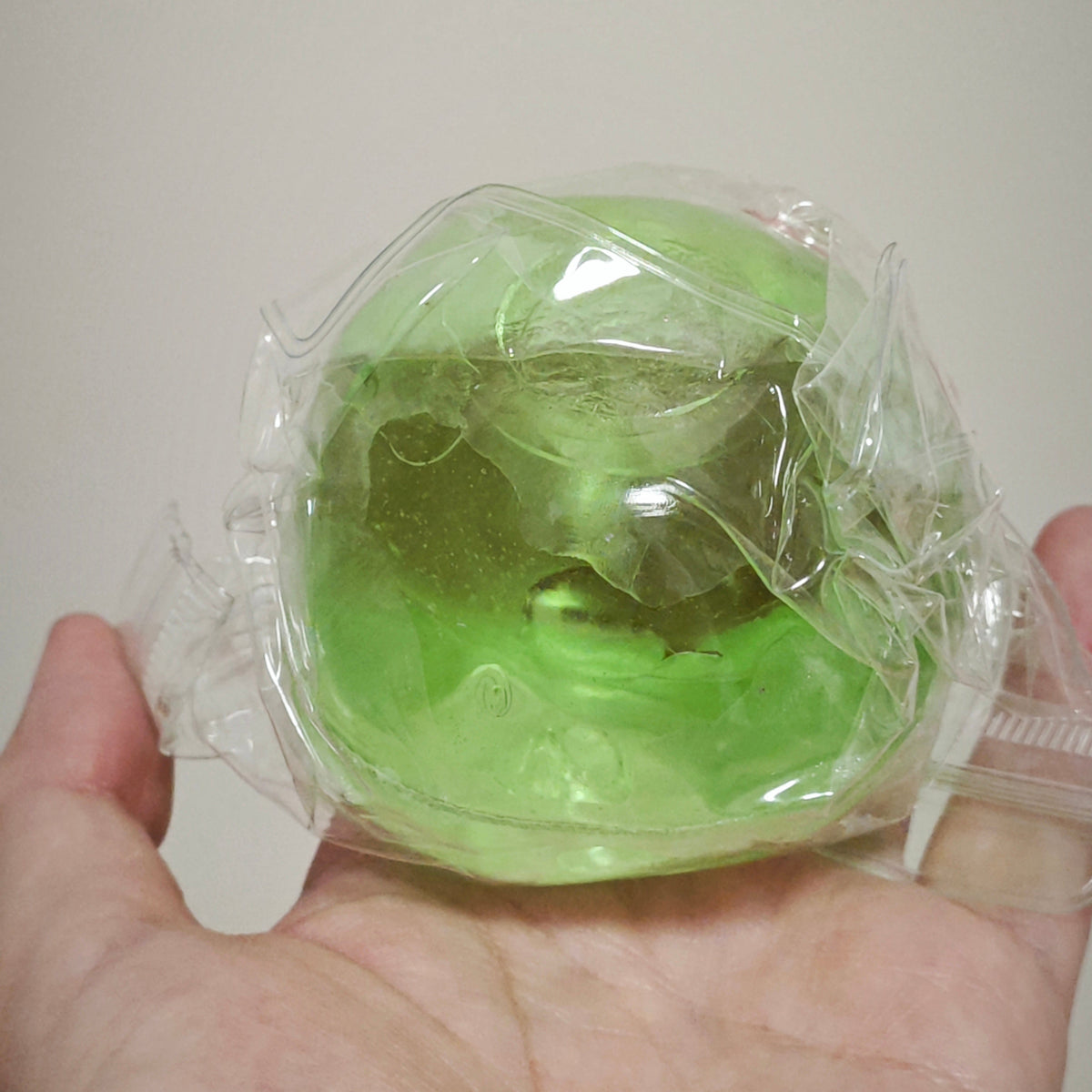 A hand holding a small, round, translucent green object wrapped in clear plastic against a plain light background.