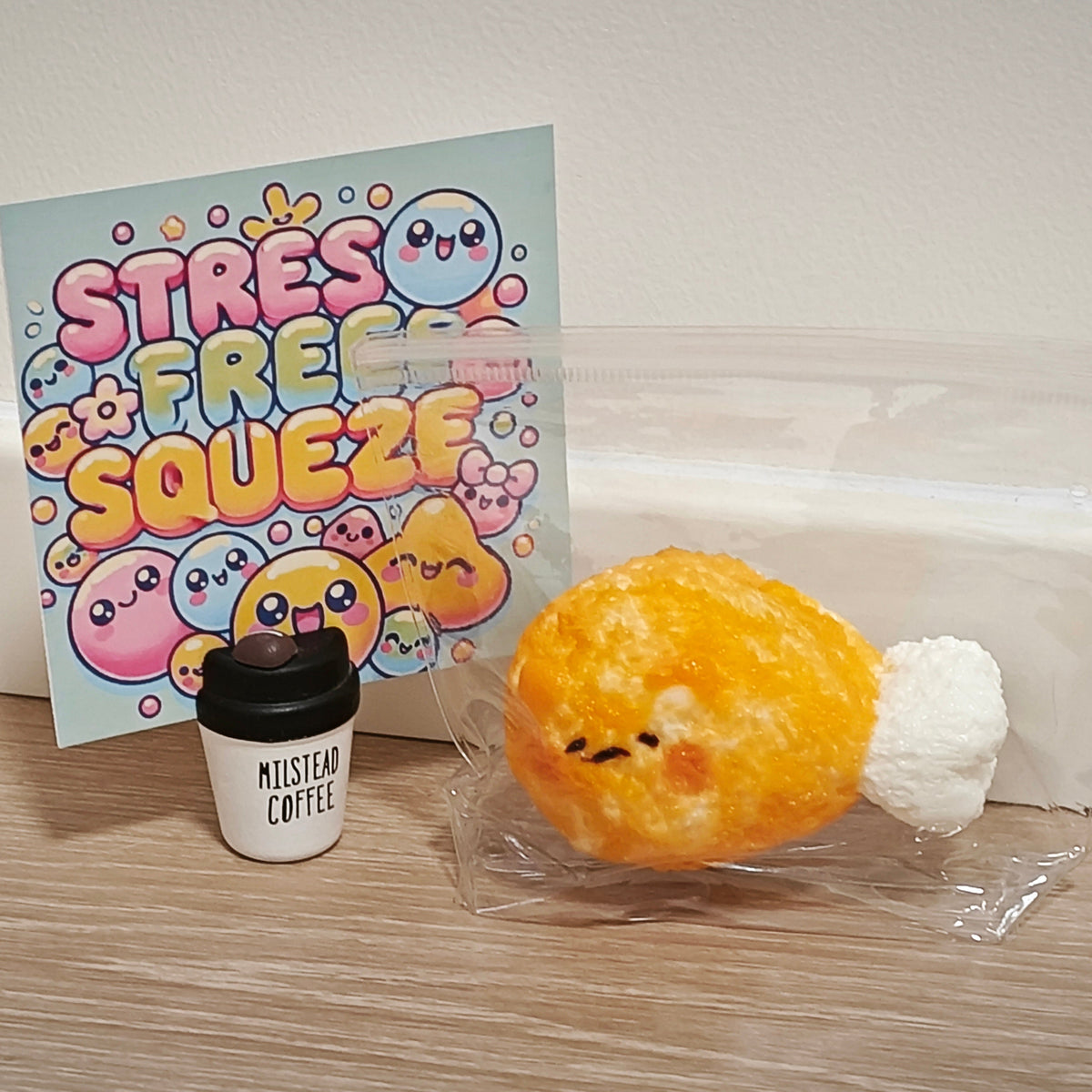 A cute yellow chicken-shaped squishy toy with a white tail sits in clear plastic packaging on a wooden table, next to a miniature white coffee cup and a colorful card that reads 'STRESS FREE SQUEEZE'.