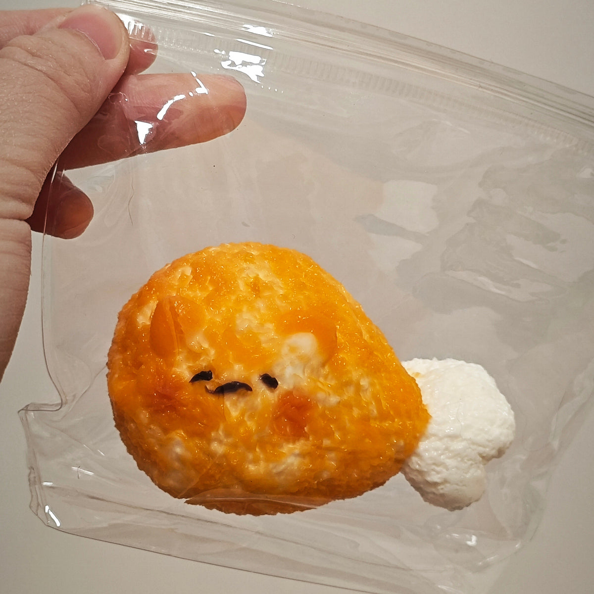 A hand holding a clear plastic zip bag containing a small plush toy shaped like a fried egg with a white fluffy tail, against a plain light background.