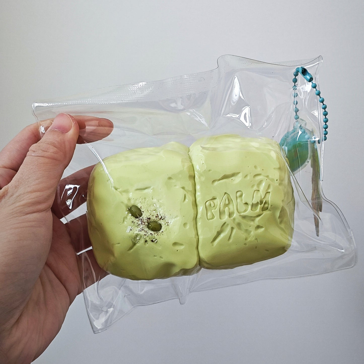 A hand holding a clear plastic bag containing two pale yellow-green squishy toys with bullet-hole details and the word 'FALKE' embossed on one piece, against a plain light gray background.