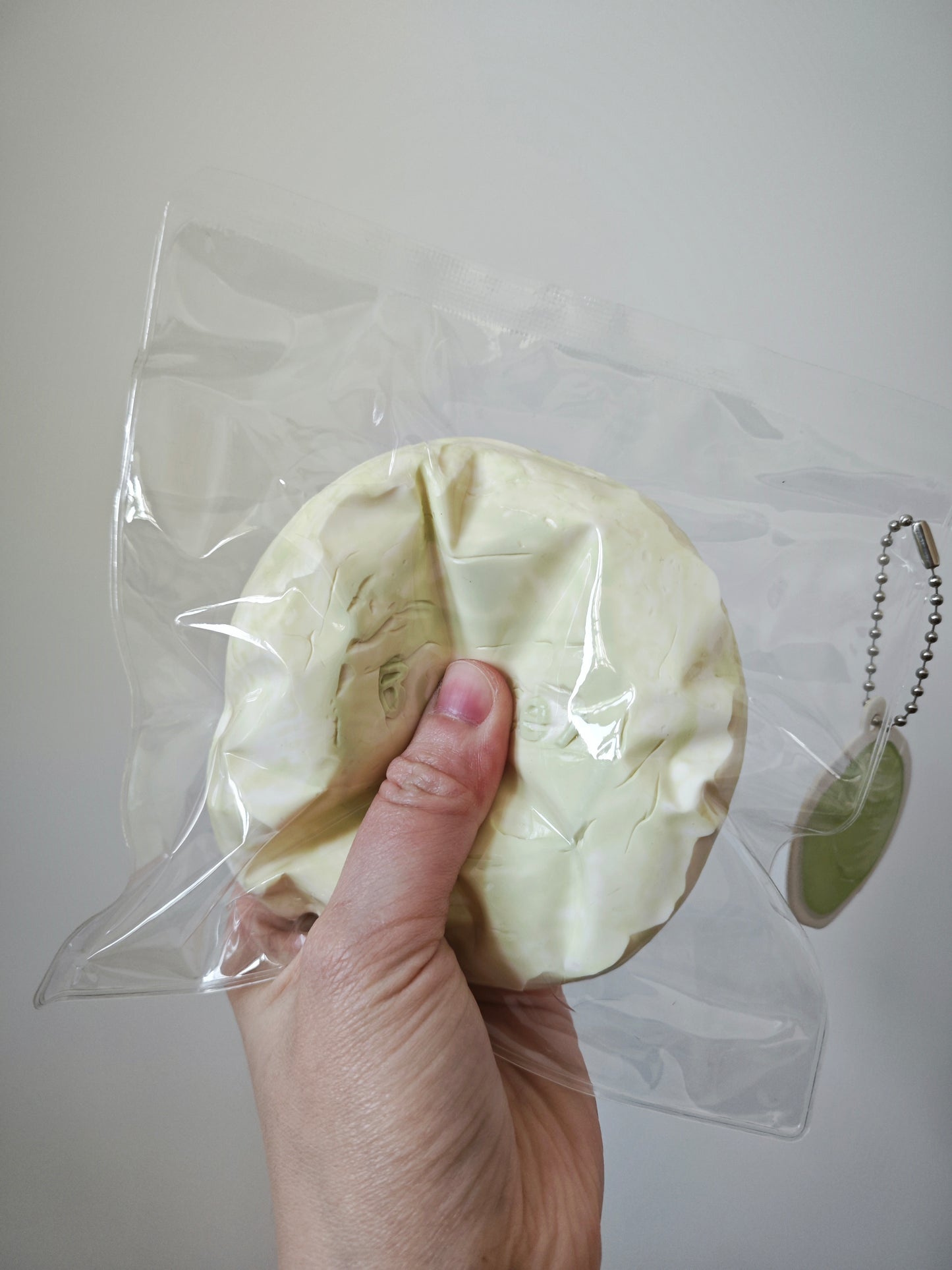 A hand holding a round, pale yellowish-beige textured item inside a clear plastic bag, with a small green oval tag attached to a silver ball chain at the right side, against a plain light gray background.