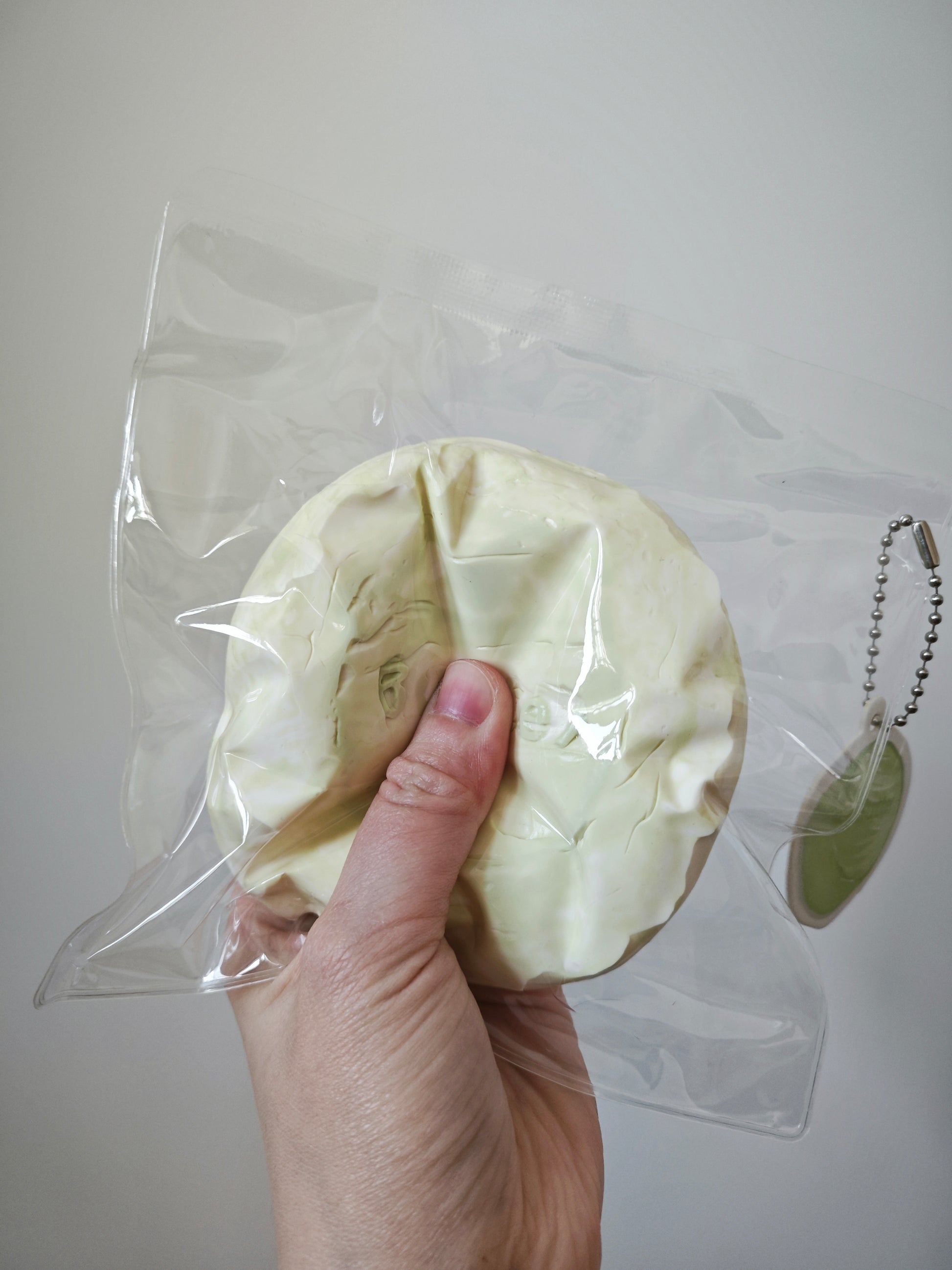 A hand holding a round, pale yellowish-beige textured item inside a clear plastic bag, with a small green oval tag attached to a silver ball chain at the right side, against a plain light gray background.