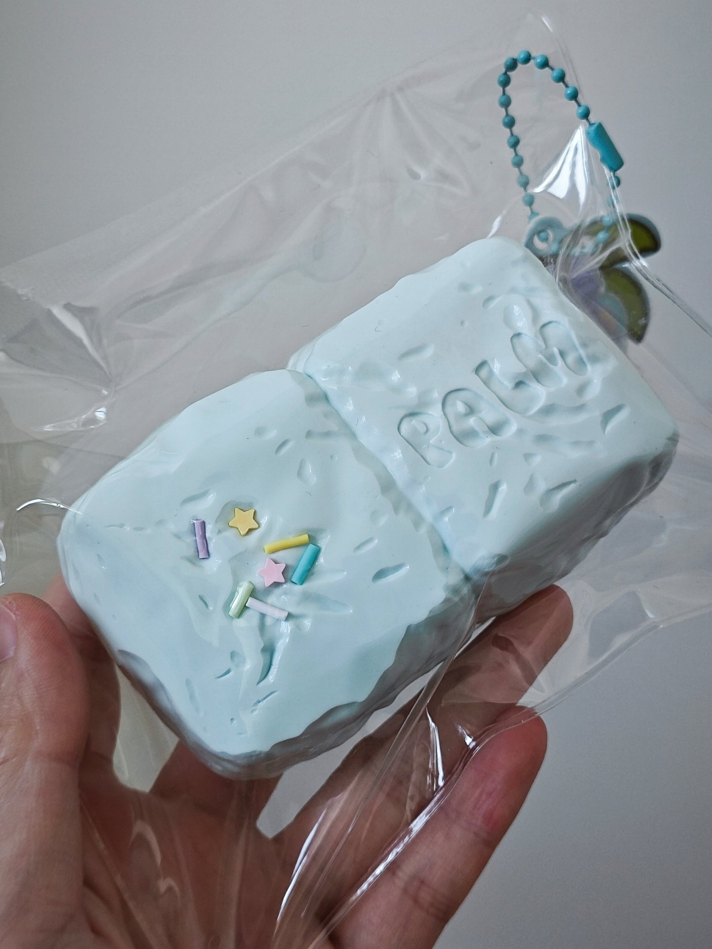 A hand holding a light blue textured squishy toy shaped like a bar of soap, wrapped in clear plastic packaging with a small blue bead chain attached.