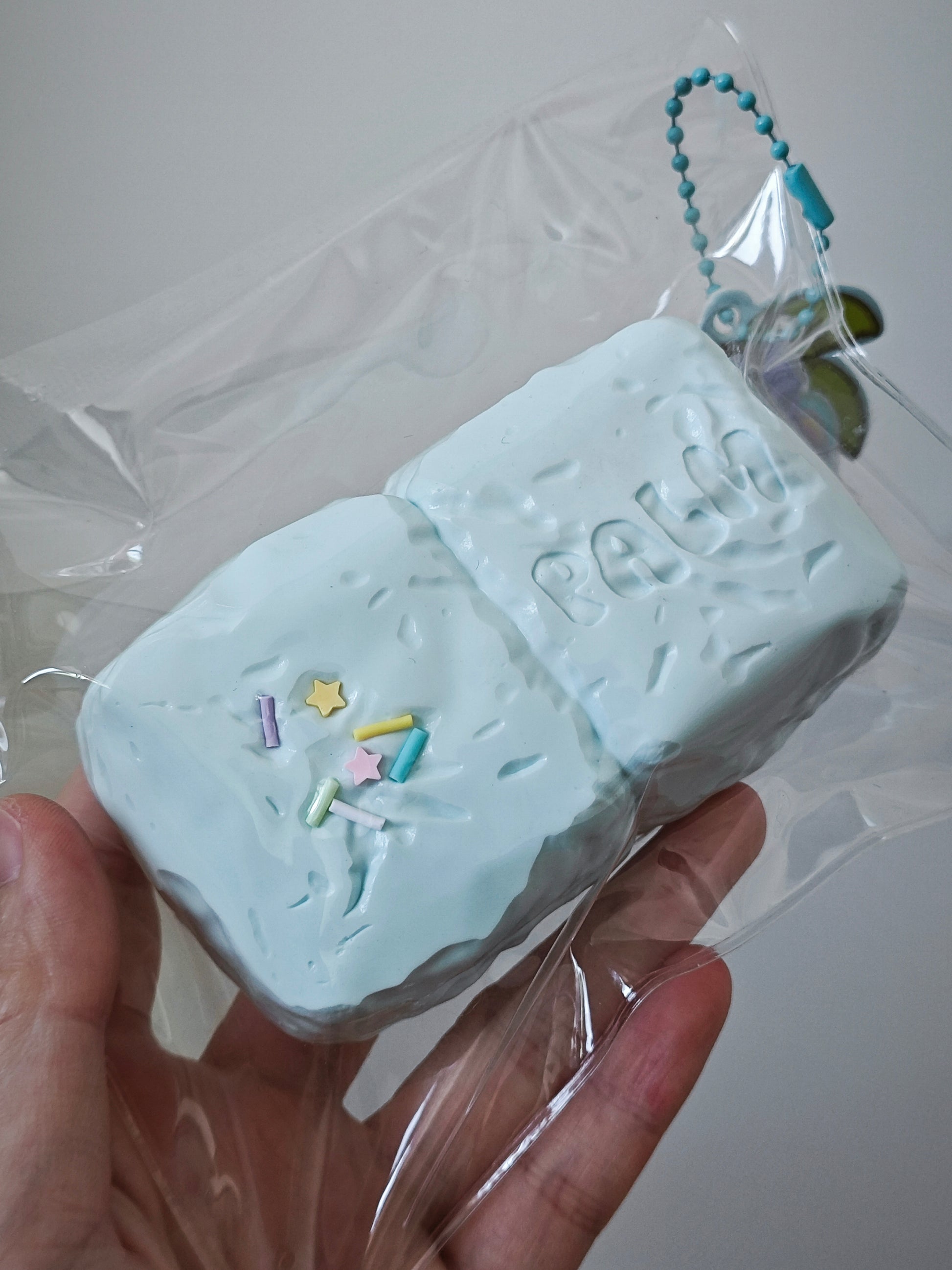 A hand holding a light blue textured squishy toy shaped like a bar of soap, wrapped in clear plastic packaging with a small blue bead chain attached.