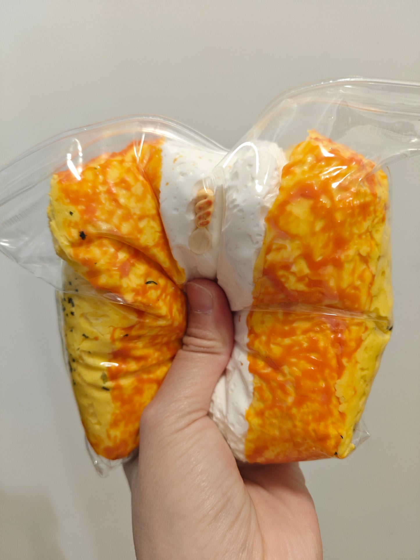 A hand holding a packaged novelty food item shaped like a split egg, with one half showing a golden-yellow scrambled egg texture and the other half showing a white creamy texture, both sealed in a clear plastic bag.
