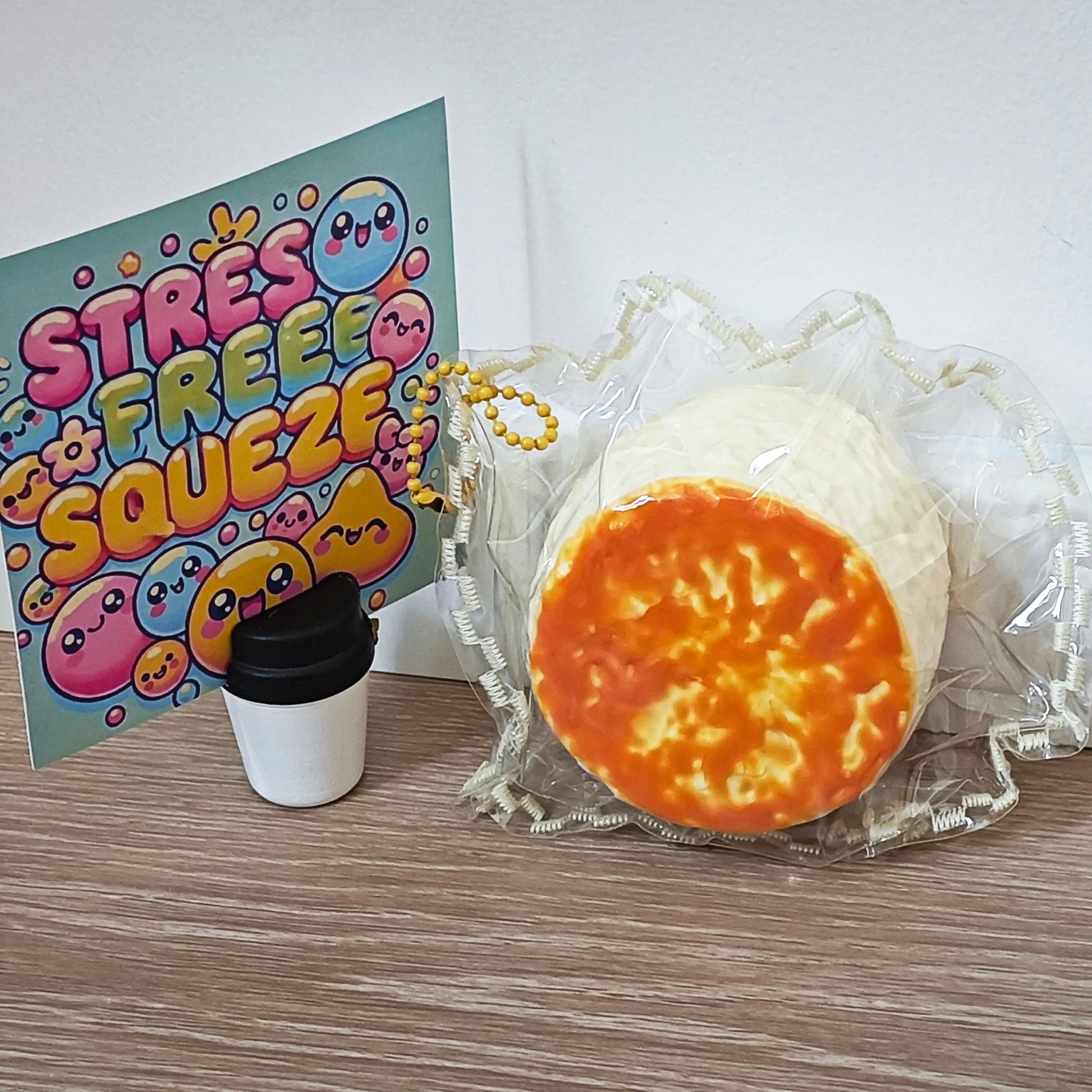 A squishy toy shaped like a slice of pizza or a baked good with a white outer layer and orange-yellow center, displayed on a wooden surface next to a small coffee cup and a colorful card that reads 'STRESS FREE SQUEEZE'.
