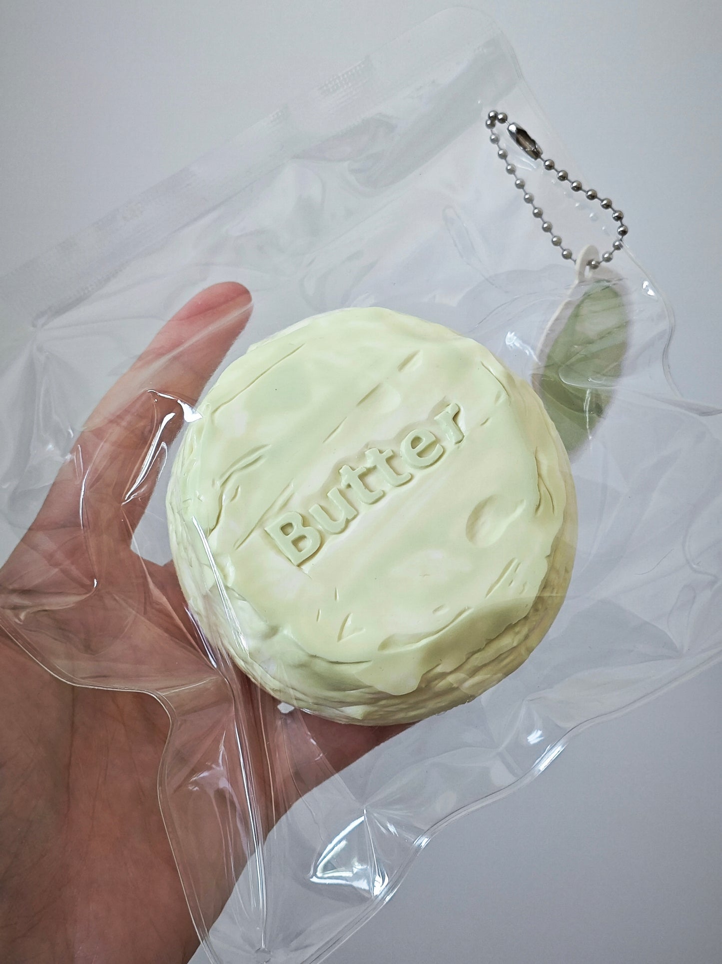 A hand holding a round, pale yellow novelty item embossed with the word 'Butter' and attached to a clear plastic bag with a metal keychain, against a plain light background.