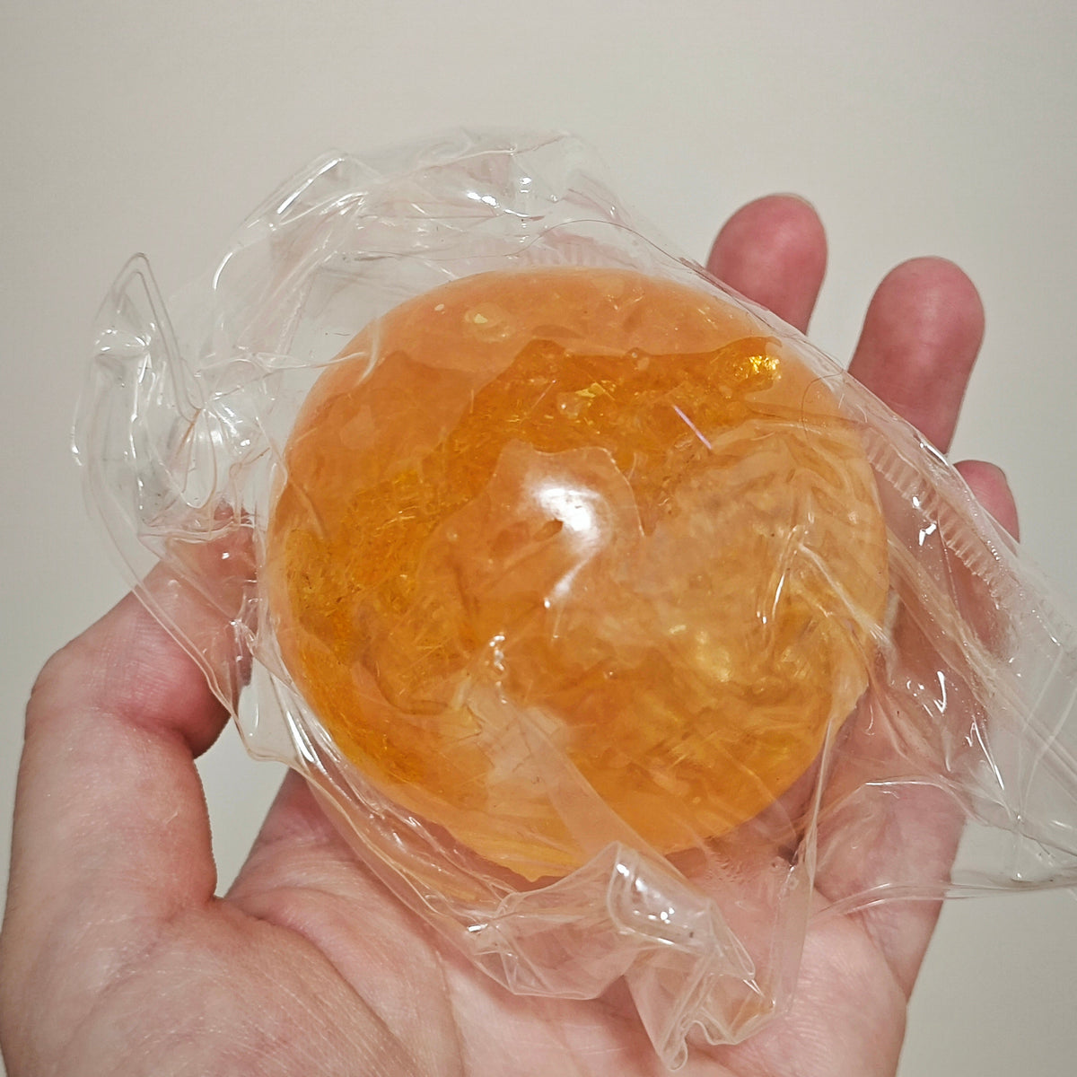 A hand holding a translucent orange gel-like sphere sealed in clear plastic packaging against a plain light background.