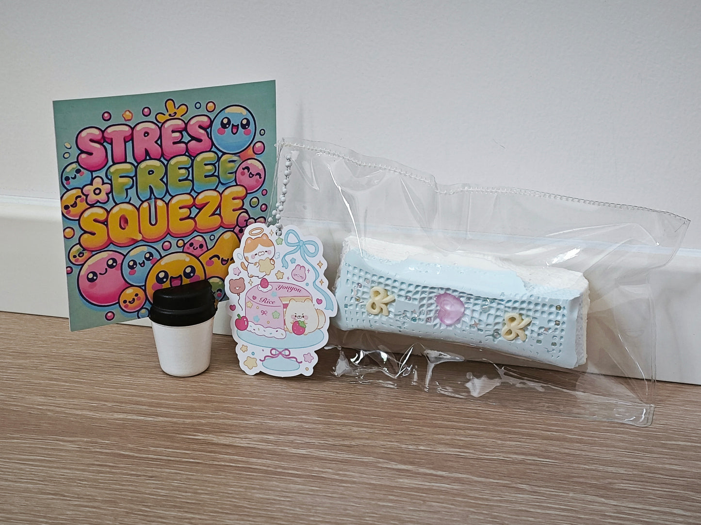 A cute stress-relief gift set on a wooden tabletop against a white wall, featuring a colorful 'STRESS FREE SQUEEZE' card, a small coffee cup, a sticker, and a light blue knitted pouch in clear plastic packaging.