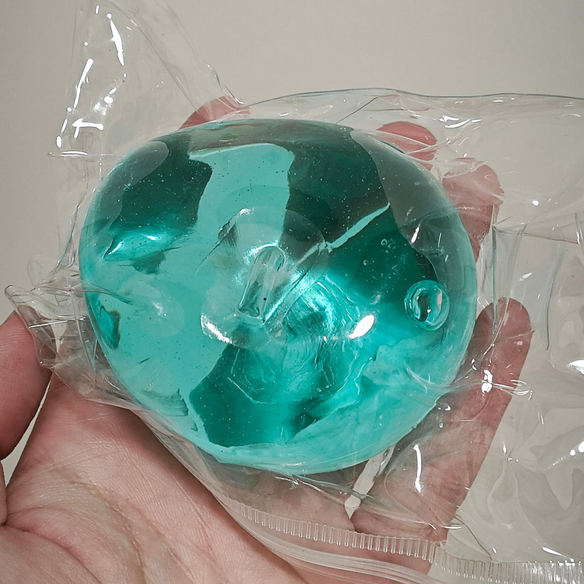 A translucent teal-green gel-like sphere sealed inside a clear plastic bag, held in a person's hand against a plain light background.