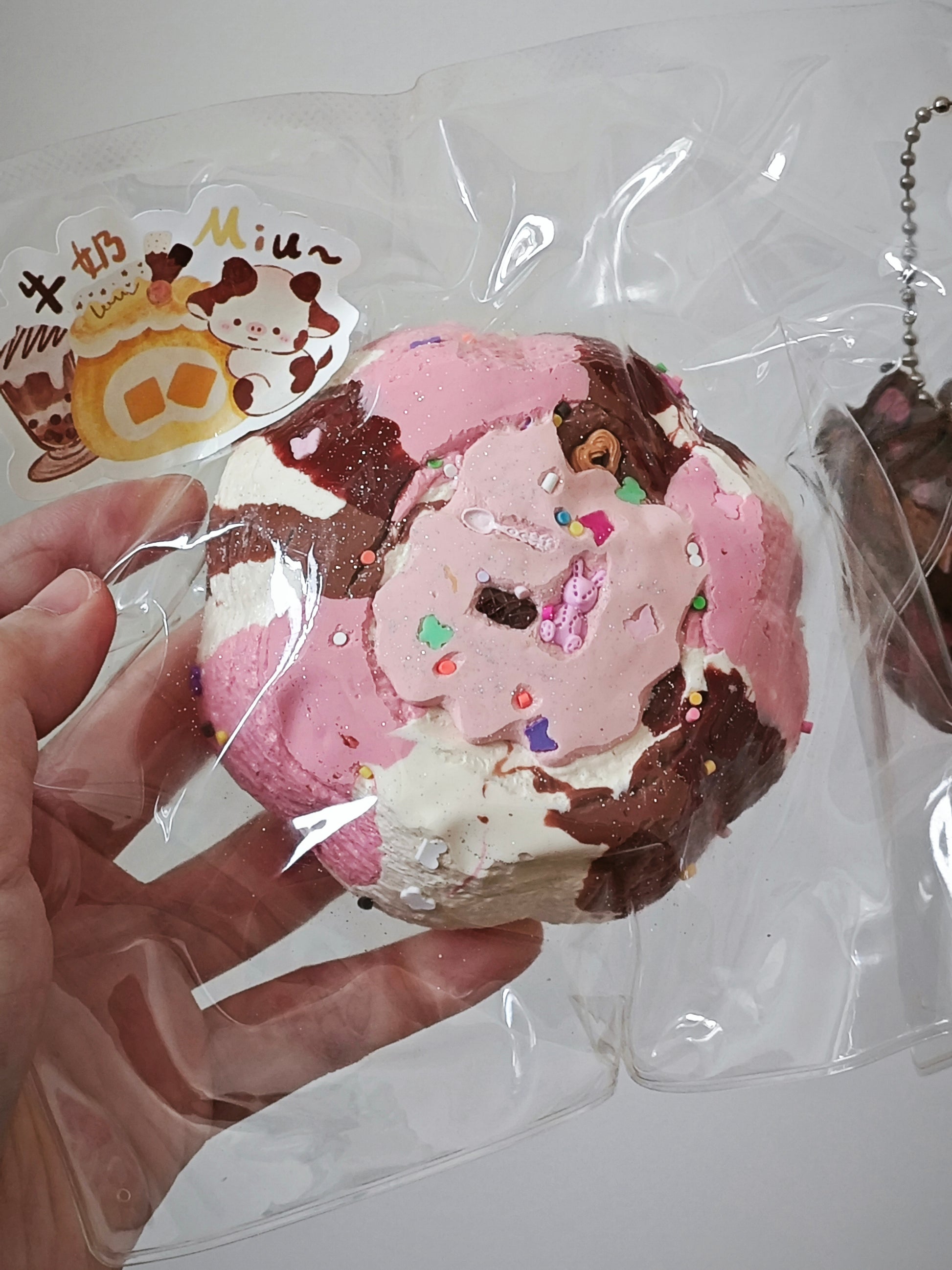A hand holds a packaged squishy toy shaped like a donut or pastry, with pink frosting, brown chocolate swirls, white cream areas, and small multicolored sprinkle details. The item is sealed in a clear plastic bag with a cartoon sticker and a metal keychain chain visible.