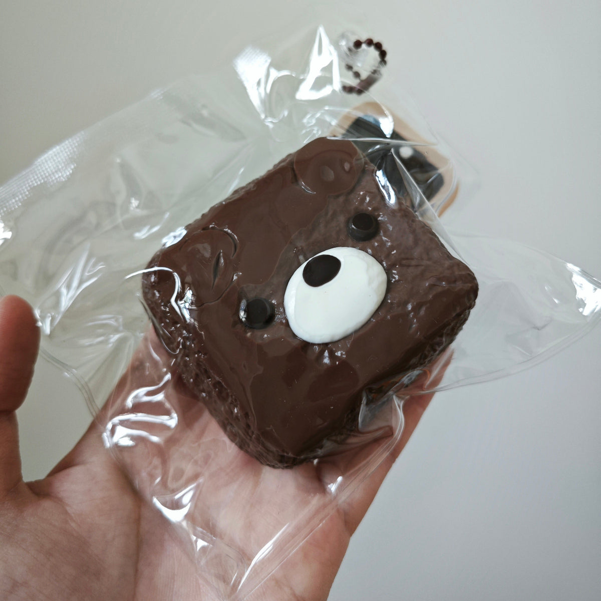 A hand holding a small square brown cake or pastry wrapped in clear plastic, decorated with a white oval face and black eyes.