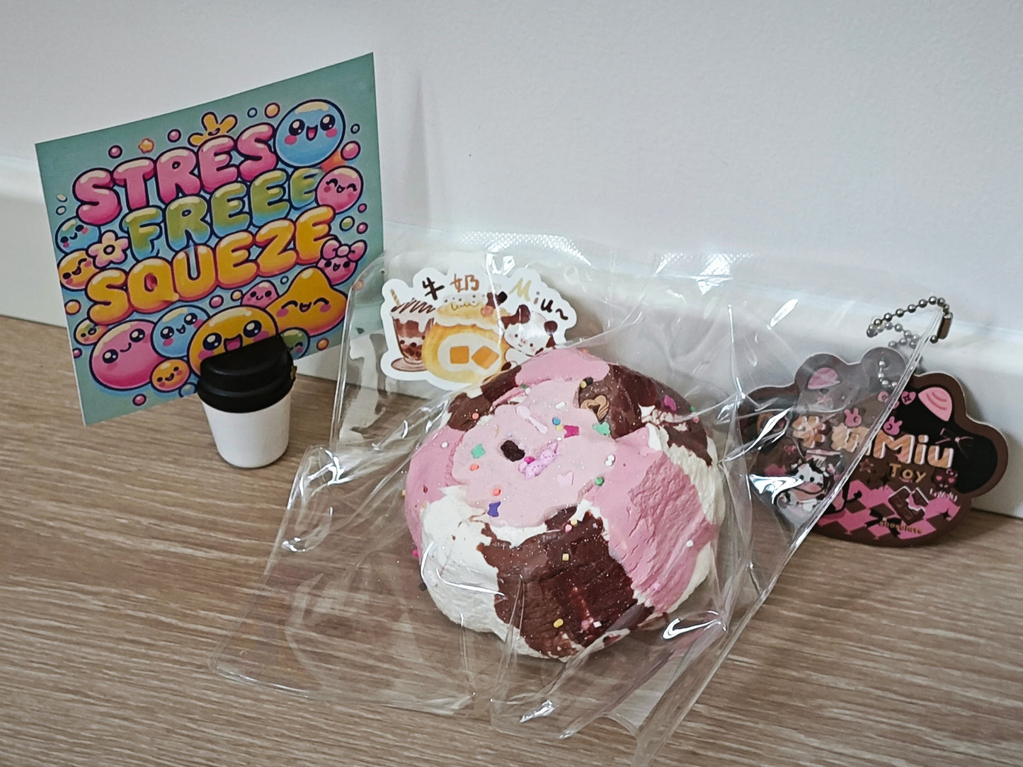 A pink and brown ice cream sundae-themed squishy toy in clear plastic packaging, displayed on a wooden surface alongside a colorful 'Stress Free Squeeze' card, a small coffee cup, a small character card, and a keychain.