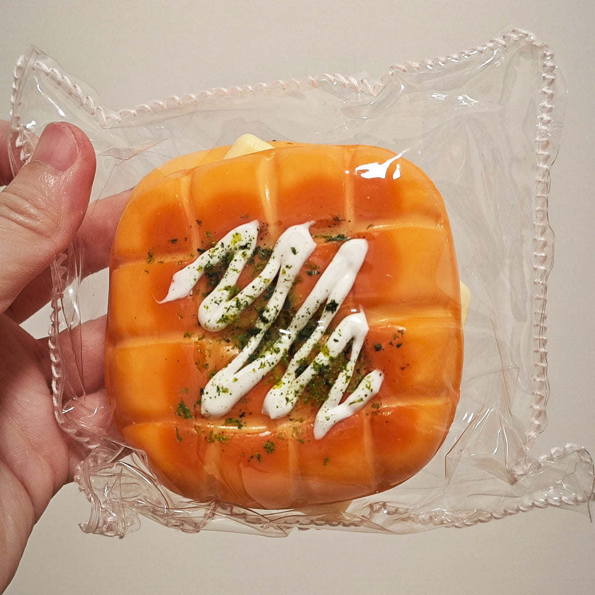 A hand holding a packaged orange-brown baked good, likely a novelty cake or pastry, topped with white drizzle and green herb flakes, sealed in clear plastic against a plain light background.