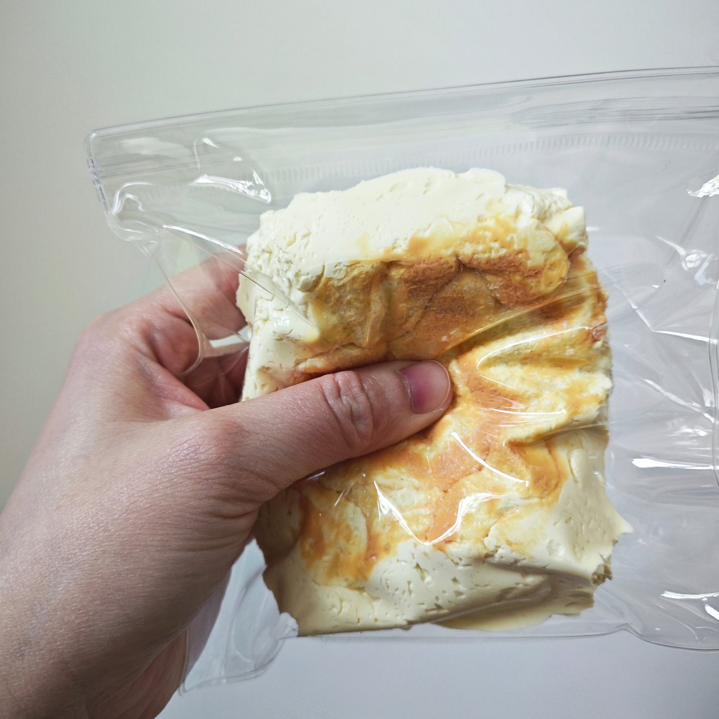 A hand holding a clear plastic bag containing a textured, crumpled foam or clay-like product in off-white and tan tones against a plain light background.