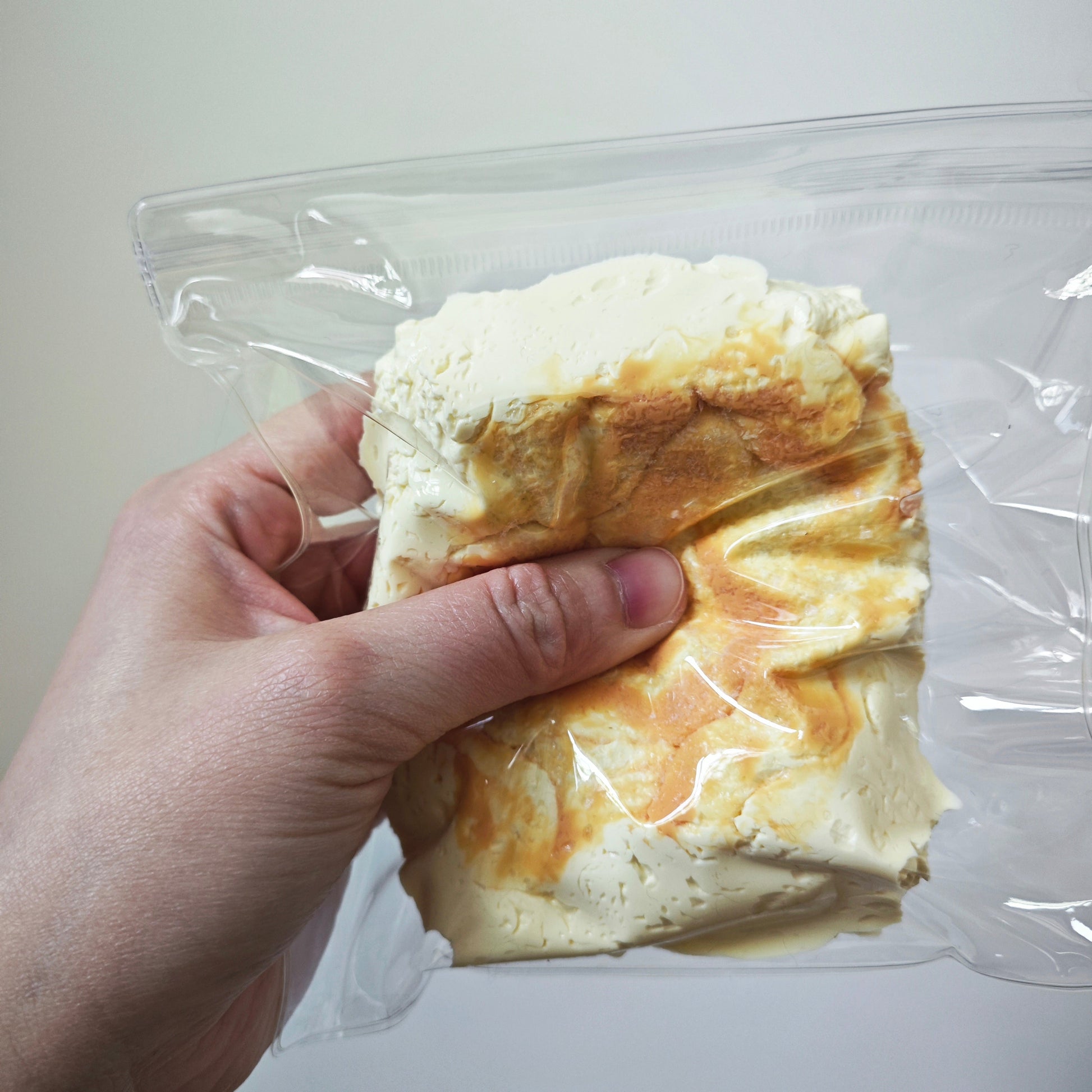 A hand holding a clear plastic bag containing a textured, crumpled foam or clay-like product in off-white and tan tones against a plain light background.