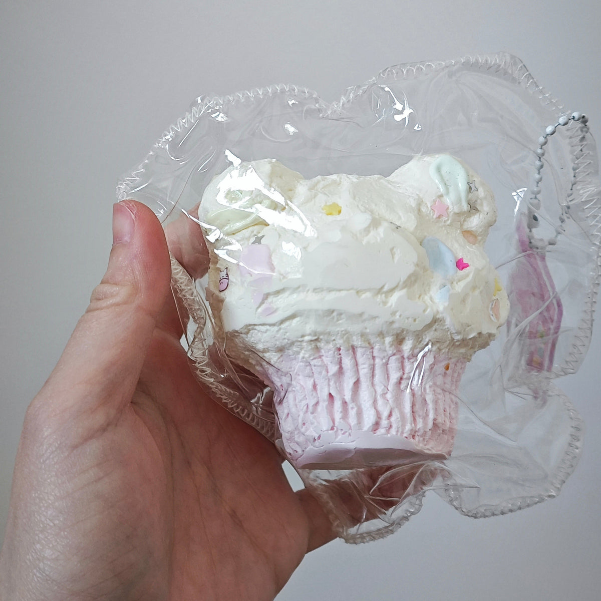 A hand holding a cupcake-shaped squishy toy wrapped in clear plastic, featuring white frosting with pastel sprinkles and a pink cupcake liner, against a plain light gray background.