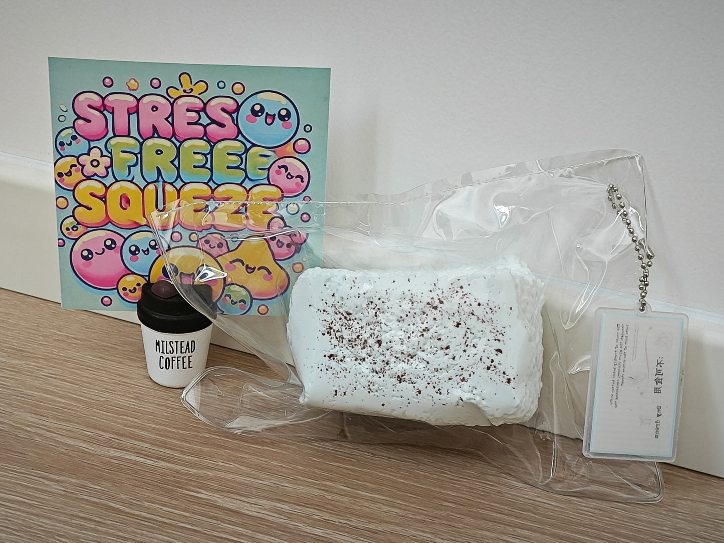 A packaged white squishy toy with dark speckles sits on a wood surface next to a small black-and-white cup labeled 'MISTLETOE COFFEE' and a colorful 'STRESS FREE SQUEEZE' card against a plain wall.