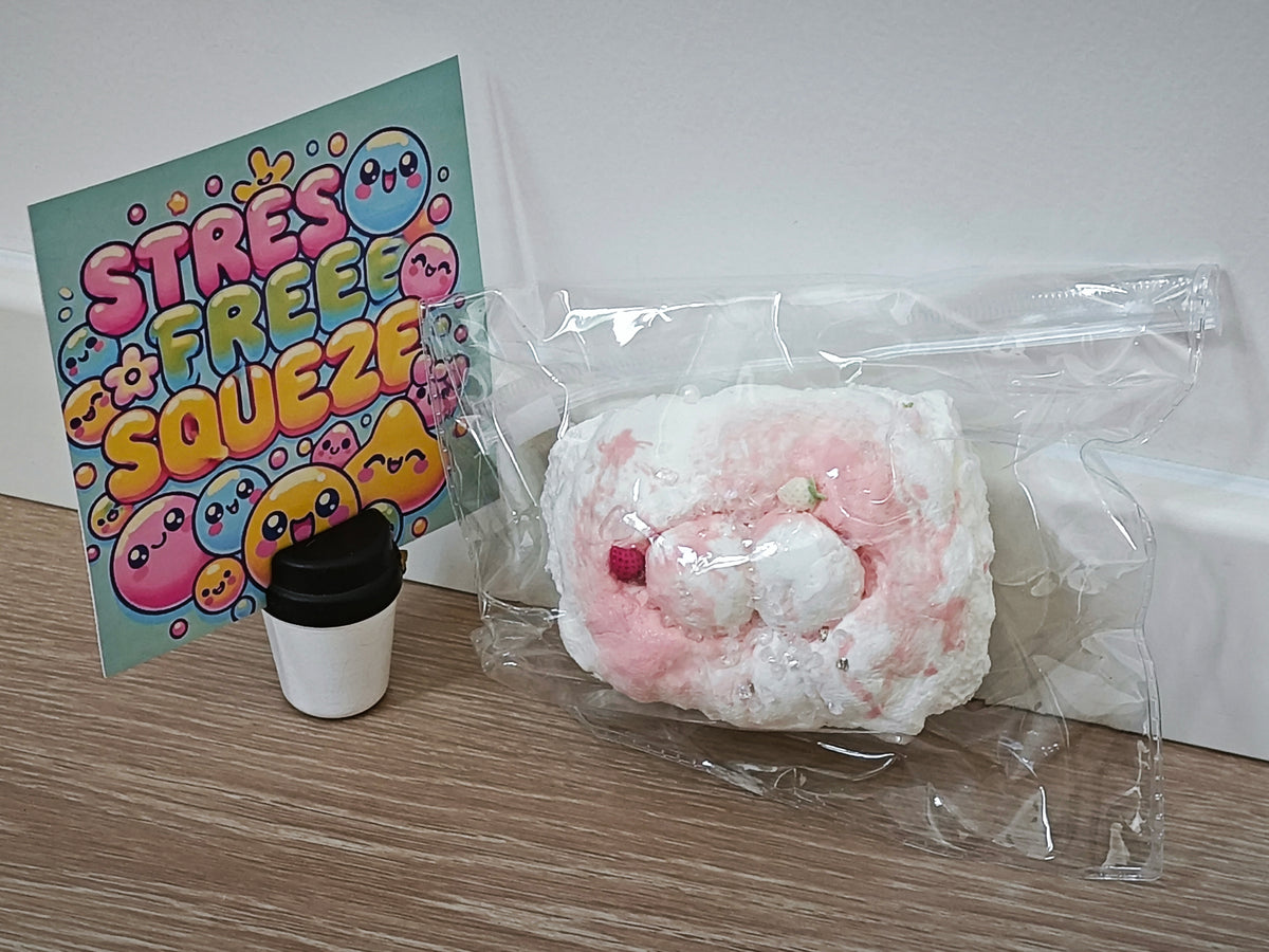 A pink and white textured stress ball sealed in a clear plastic bag, displayed next to a small black-and-white coffee cup and a colorful 'Stress Free Squeeze' card on a wooden tabletop against a white wall.