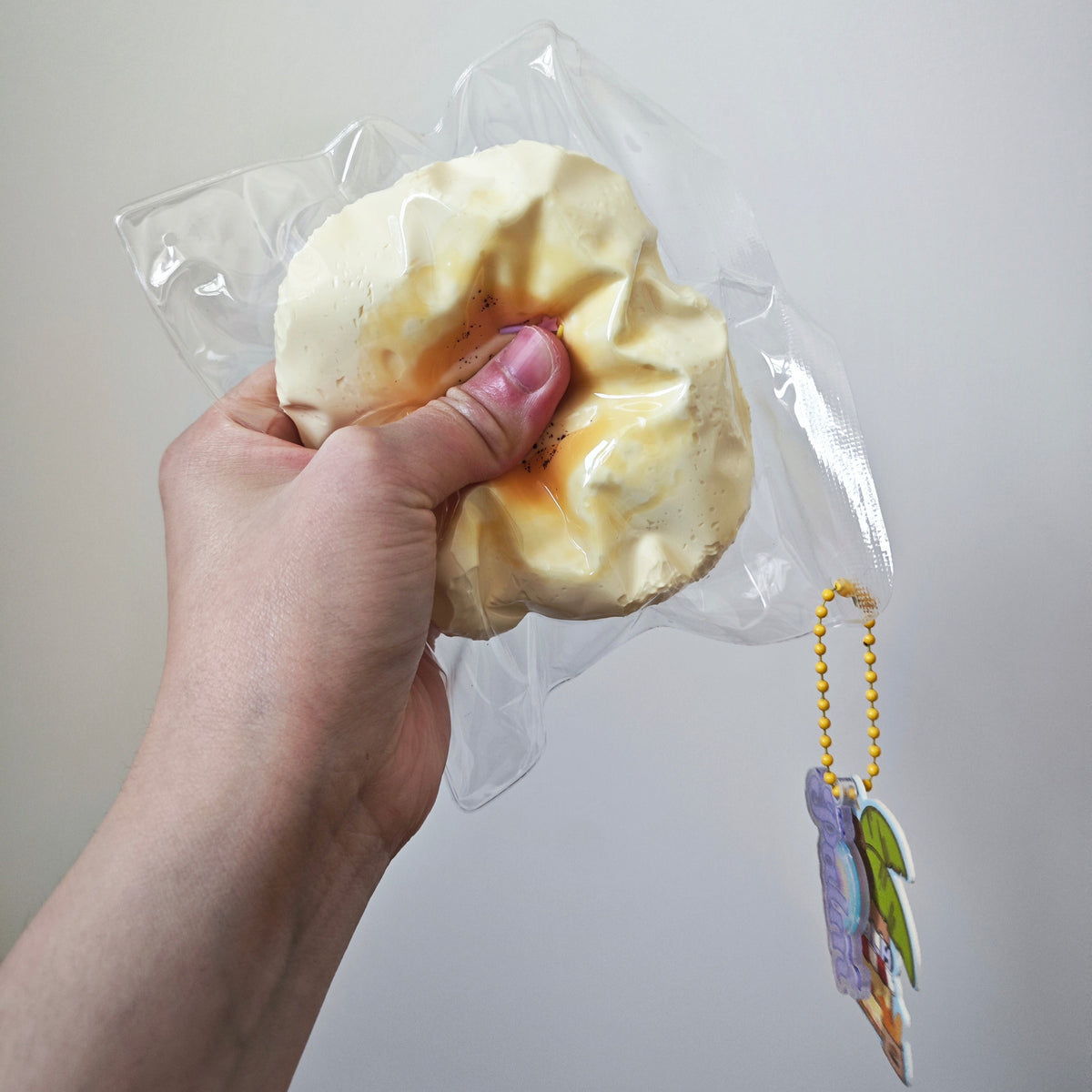 A hand holding a packaged squishy toy shaped like a burnt bread roll, with a small hanging charm attached, against a plain light gray background.