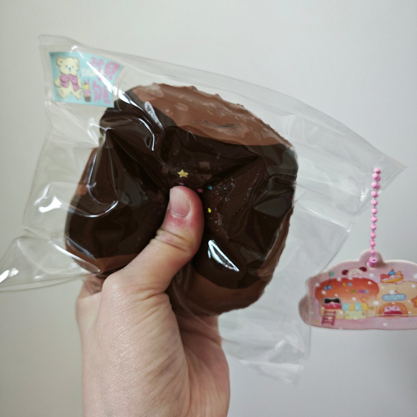 A hand holding a chocolate-donut-shaped squishy toy inside a clear plastic bag, with a small pink character charm visible on the right against a plain light background.