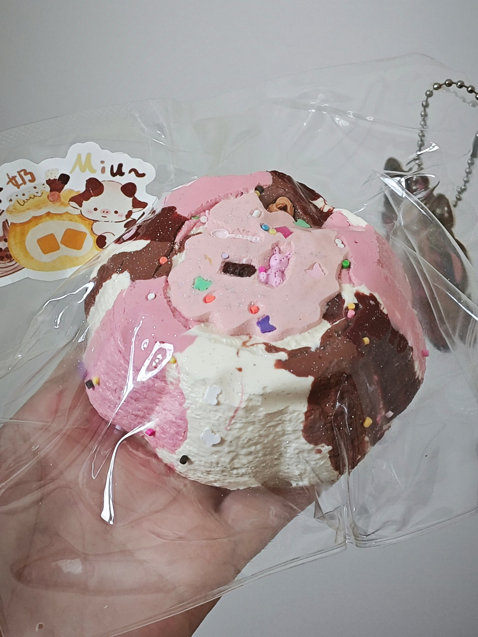 A hand holds a pastel pink and white donut-shaped squishy toy with brown chocolate drizzle and colorful sprinkles, wrapped in clear plastic packaging with a decorative sticker and chain visible.