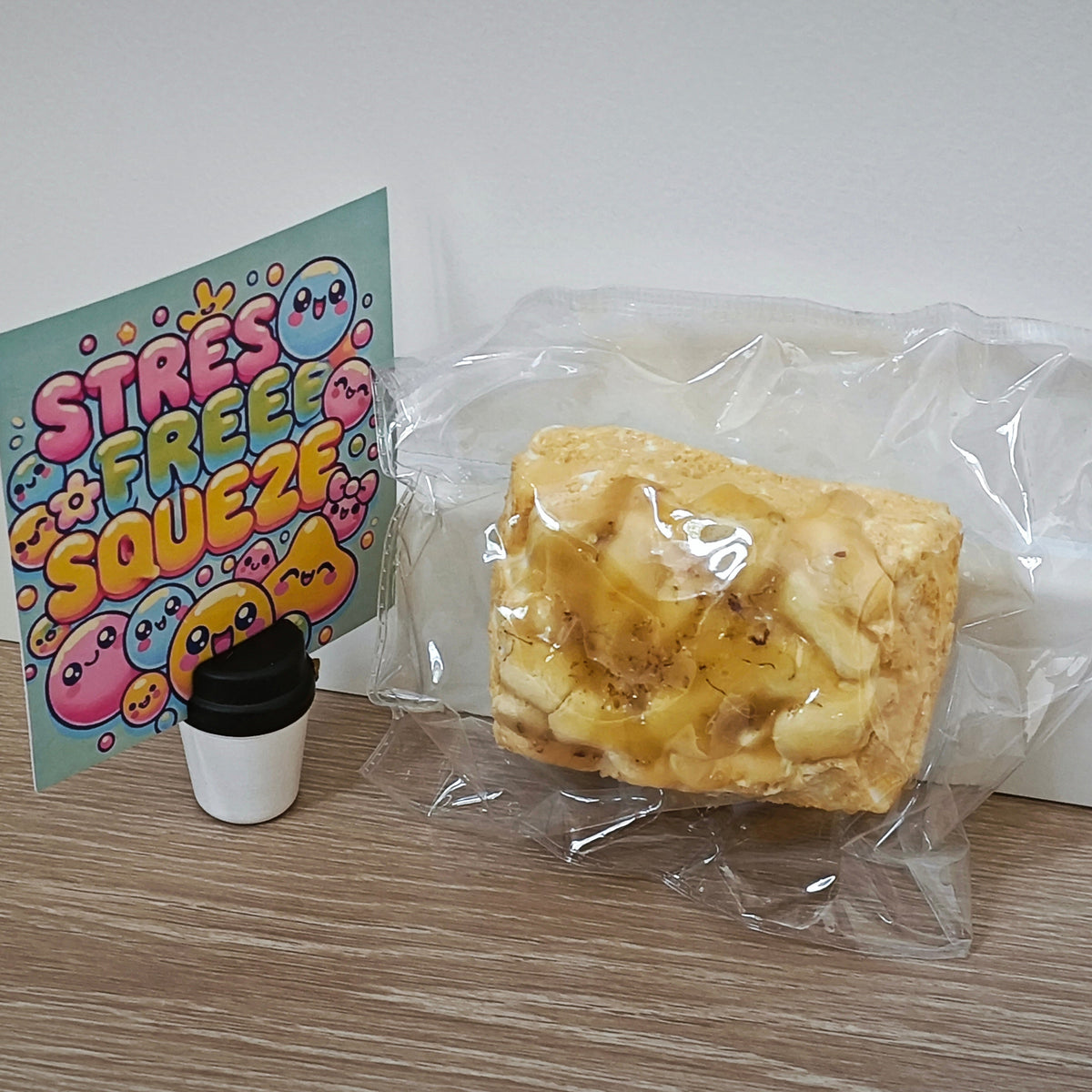 A packaged yellow cake-like squishy toy sits on a wood floor against a white wall, next to a small white cup with a black lid and a colorful card that reads 'STRESS FREE SQUEEZE'.