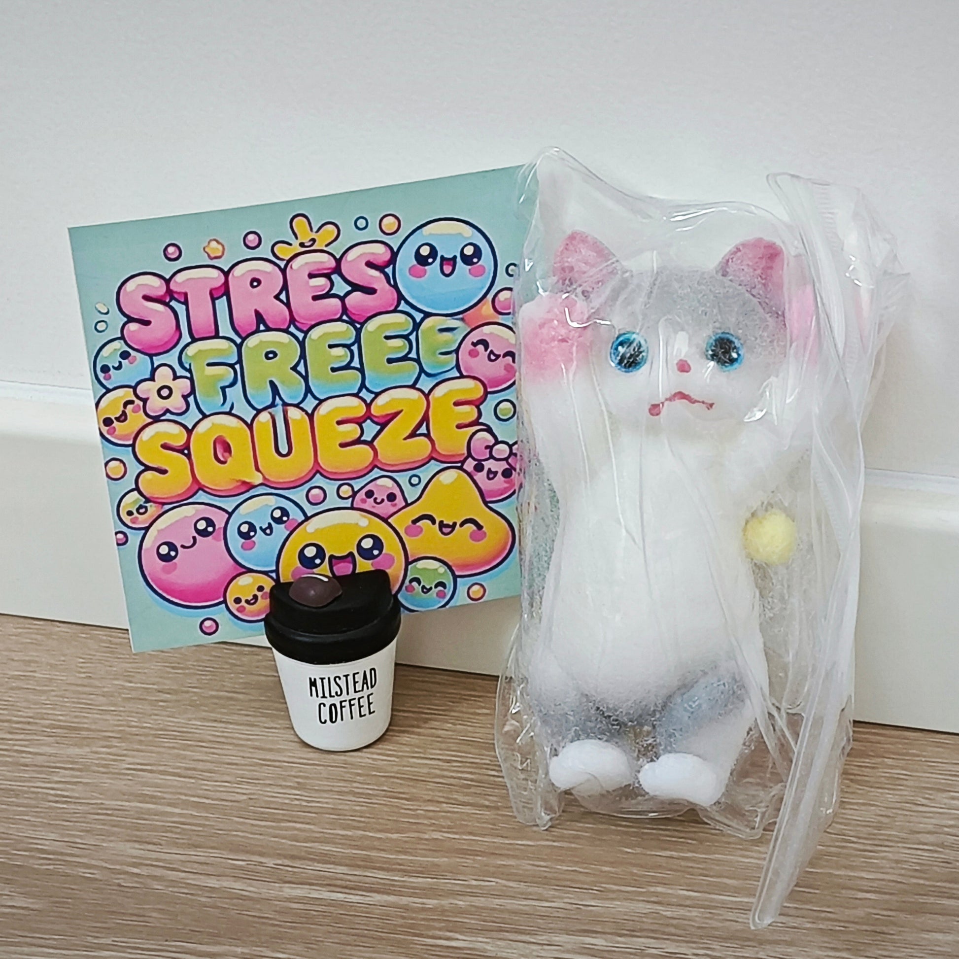 A plush white and gray cat toy sealed in a clear plastic bag sits on a wood floor next to a colorful 'Stress Free Squeeze' card and a small 'Miststeam Coffee' cup, against a white wall.