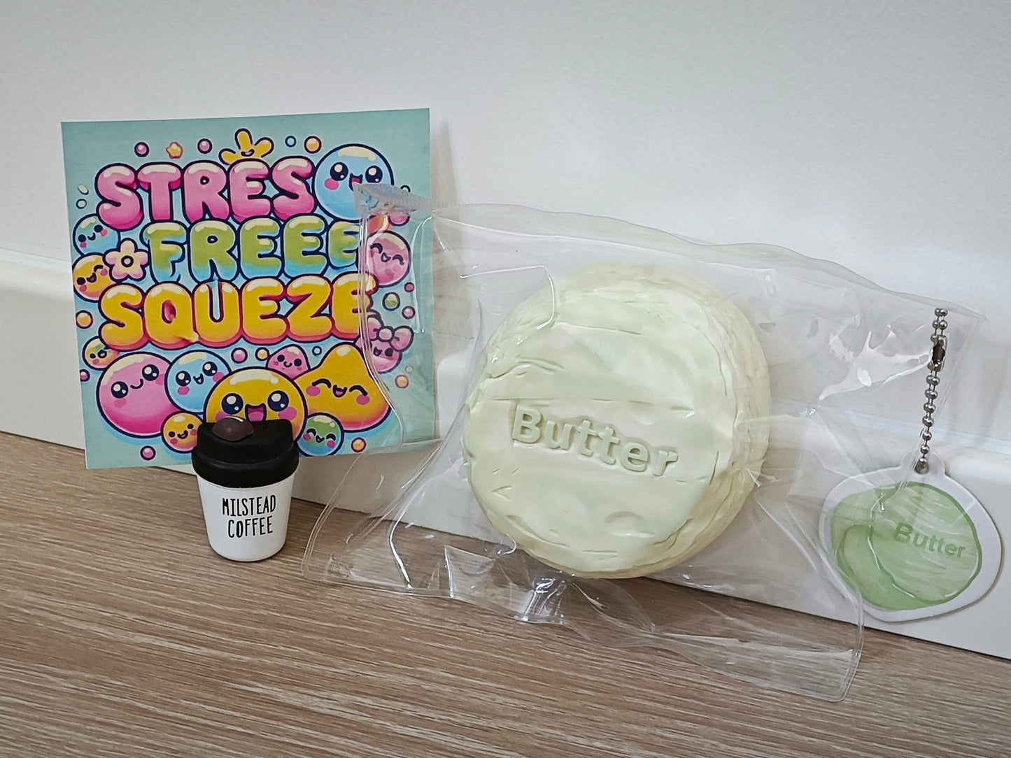 A stress-free squishy toy set on a wooden surface against a white wall, featuring a large pale yellow-green round squishy labeled 'Butter' in clear packaging, a small black-and-white coffee cup charm labeled 'MUSTARD COFFEE', and a colorful 'STRESS FREE SQUEEZE' card.
