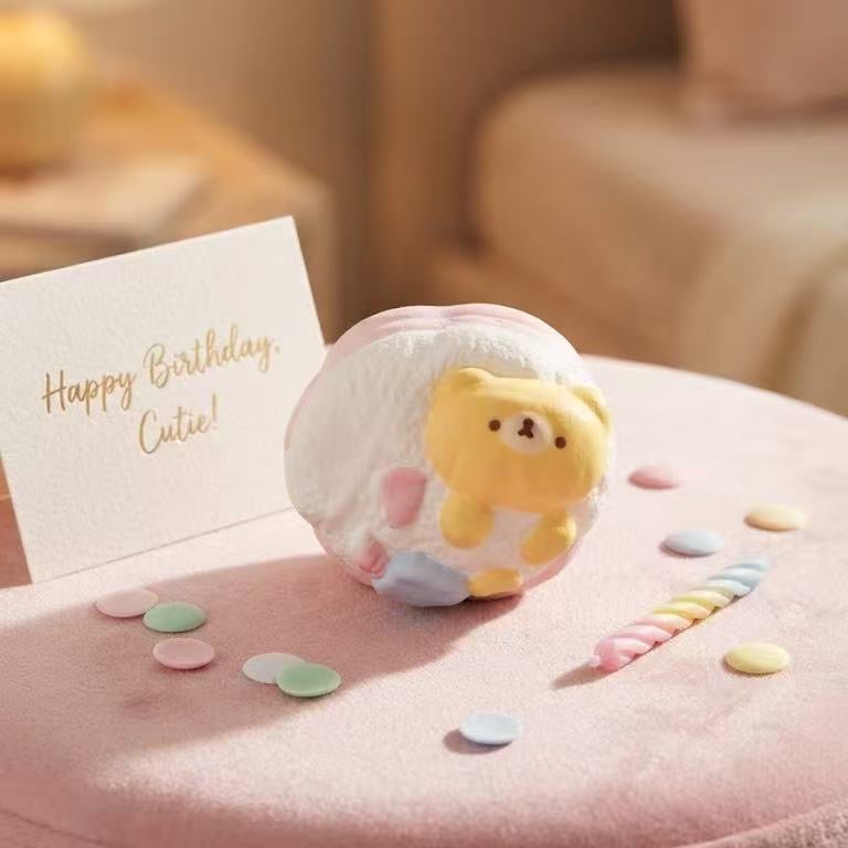 A cute pastel pink and white egg-shaped candle holder with a yellow bear face, displayed on a pink velvet ottoman next to a 'Happy Birthday, Cute!' card and pastel confetti.