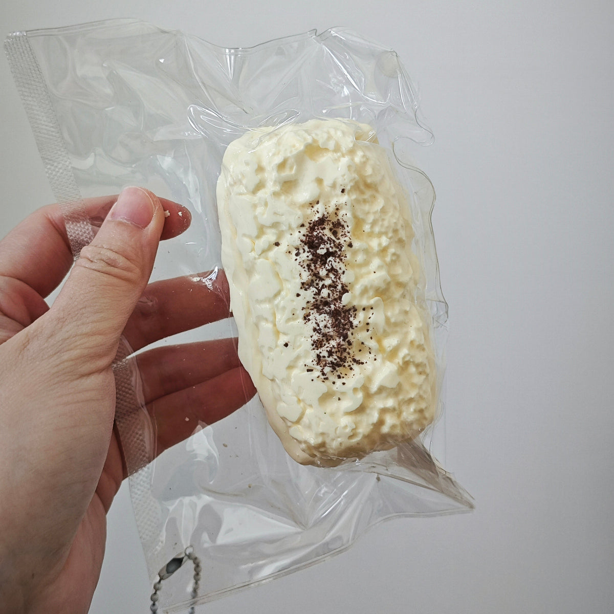 A hand holding a clear plastic bag containing a rectangular, textured off-white food item topped with a dark brown sprinkle, against a plain light background.