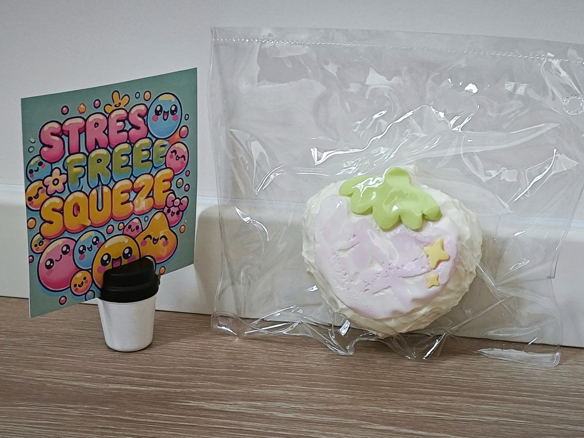 A round, sugar-dusted squishy toy with a light pink face, yellow stars, and a green leaf on top, sealed in a clear plastic bag next to a small coffee cup and a colorful 'STRESS FREE SQUEEZE' card on a wooden surface.