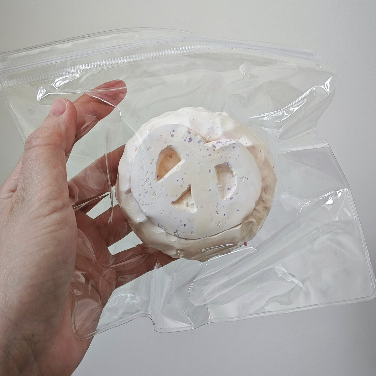 A hand holding a clear plastic zip-top bag containing a round white novelty item with a peace sign cutout and small multicolor speckles, against a plain light gray background.