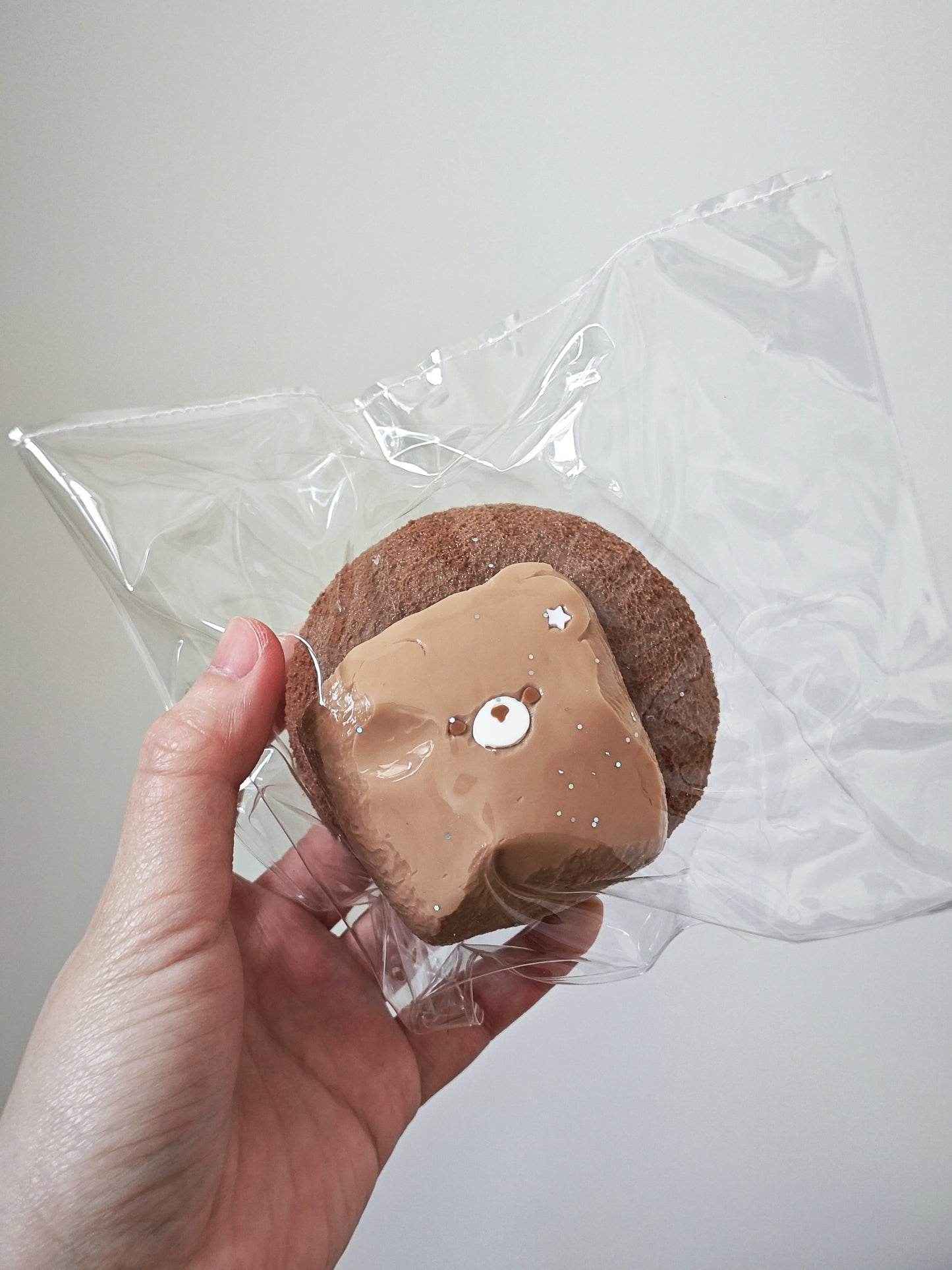 A hand holding a round brown cookie topped with a glossy light brown chocolate or caramel drizzle, both sealed in a clear plastic bag against a plain light background.