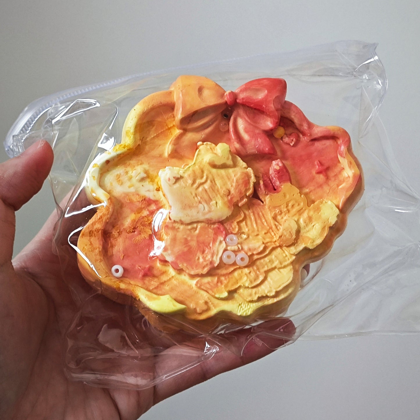 A hand holding a packaged, handcrafted cookie shaped like a stylized fish with yellow, orange, and pink icing details, sealed in a clear plastic bag against a plain light gray background.