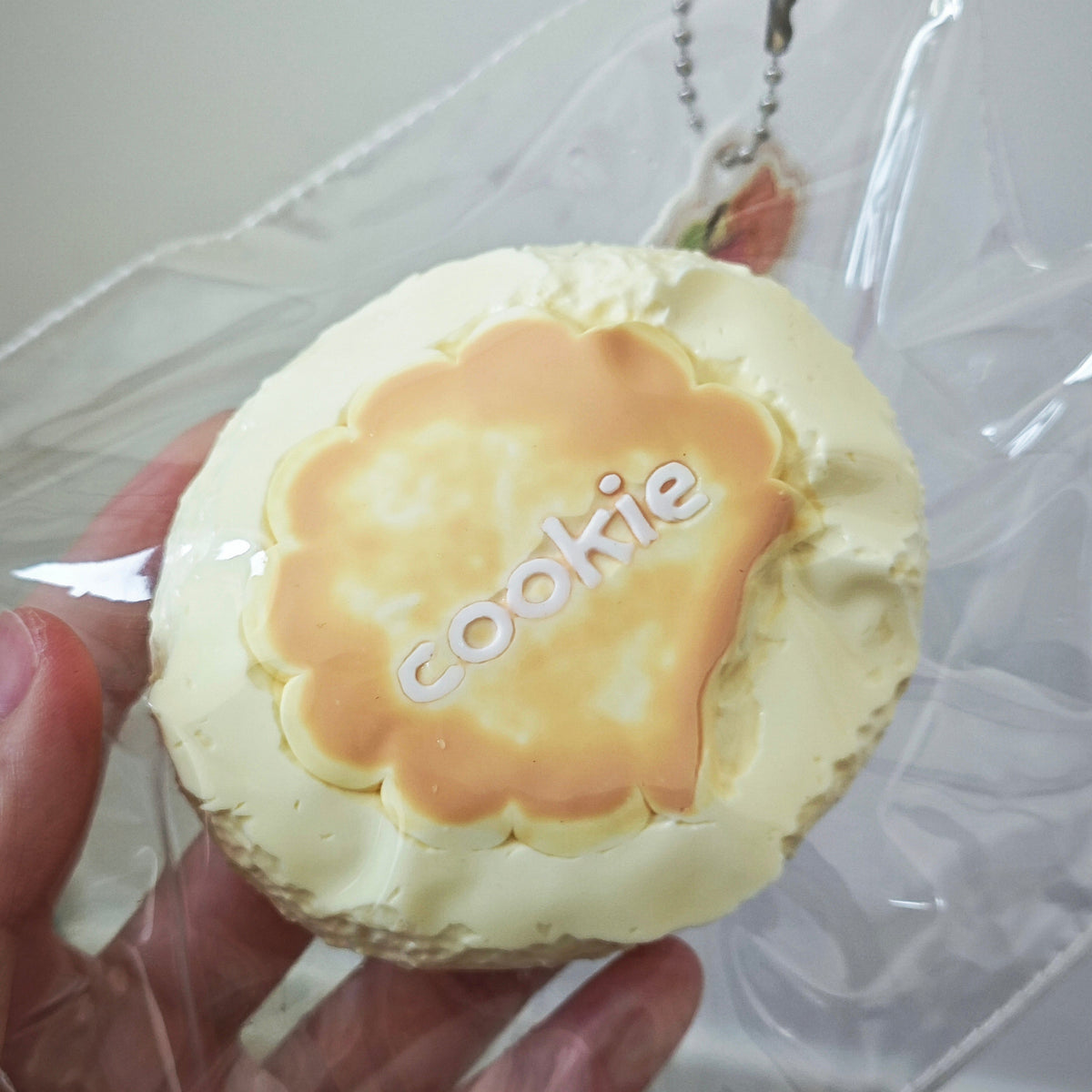 A hand holding a round, cupcake-like novelty item wrapped in clear plastic. The item has pale cream frosting around the edge and a golden-brown center with the word 'cookie' in white script.