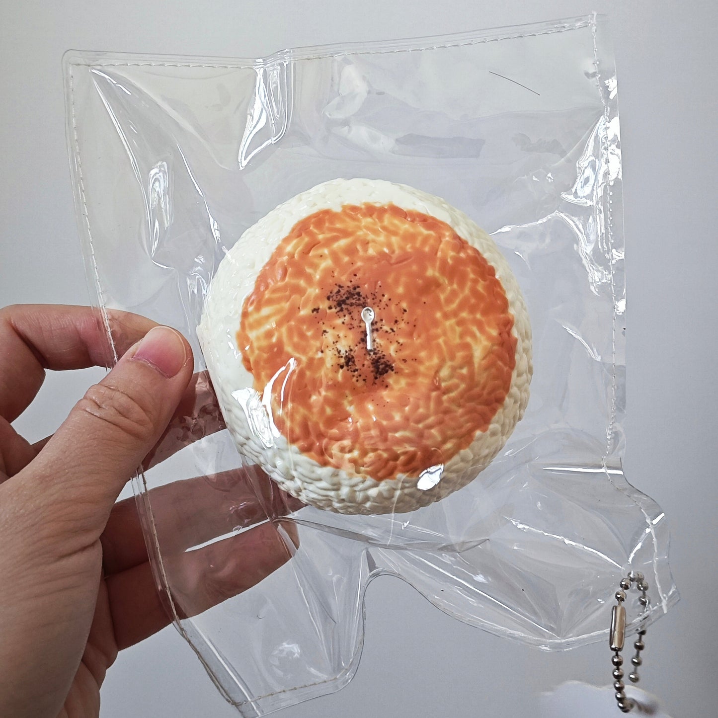 A hand holding a round, bread-like novelty item inside a clear plastic bag against a plain light gray background.