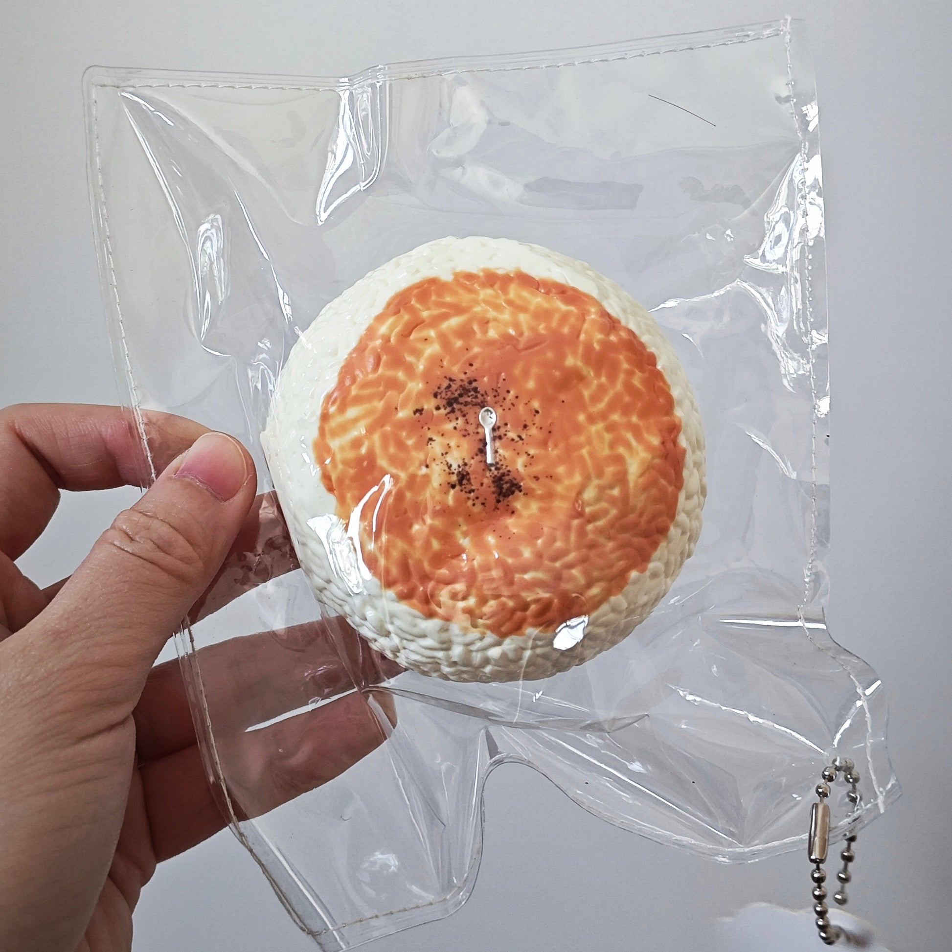 A hand holding a round, bread-like novelty item inside a clear plastic bag against a plain light gray background.
