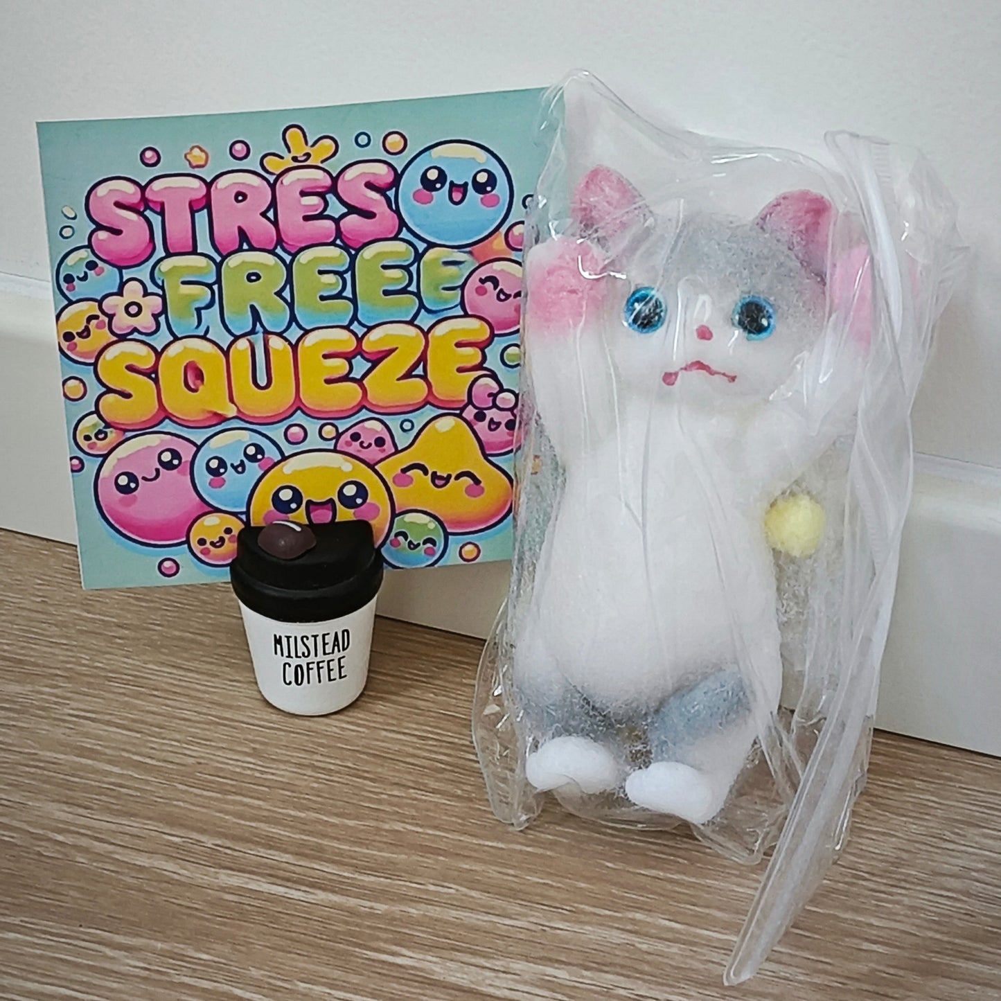 A plush cat toy wrapped in clear plastic stands next to a colorful card reading 'STRESS FREE SQUEEZE' and a small cup labeled 'MUSTSEE Coffee' on a wood floor against a white wall.