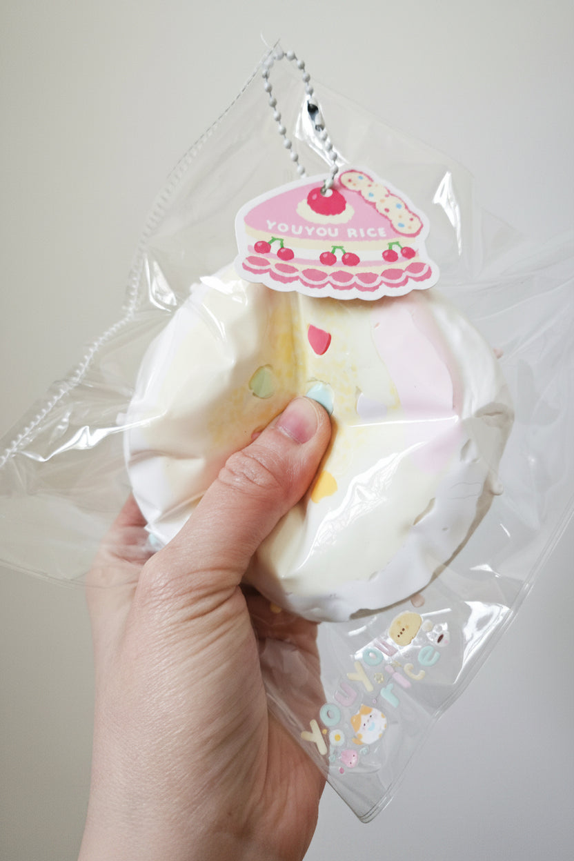 A hand holding a packaged squishy toy shaped like a slice of cake or pastry, sealed in clear plastic with a decorative hanging tag that reads 'YOU YOU RICE'.