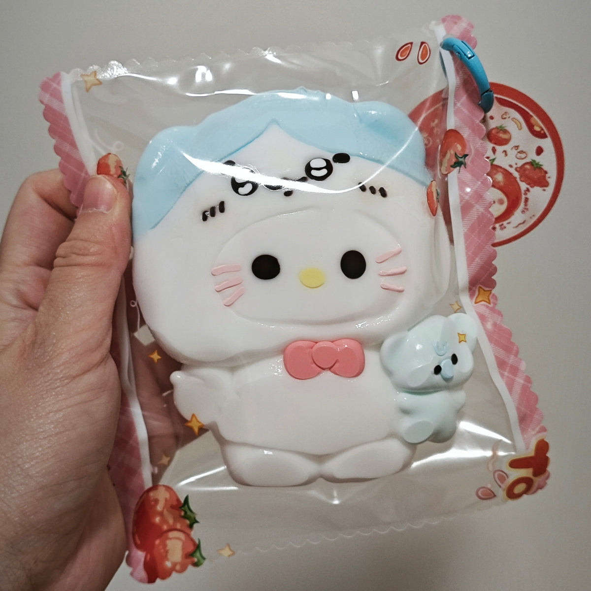 A hand holding a packaged white cartoon character toy with a light blue hat, pink bow, and small light blue animal companion inside a clear plastic bag with strawberry-themed print.