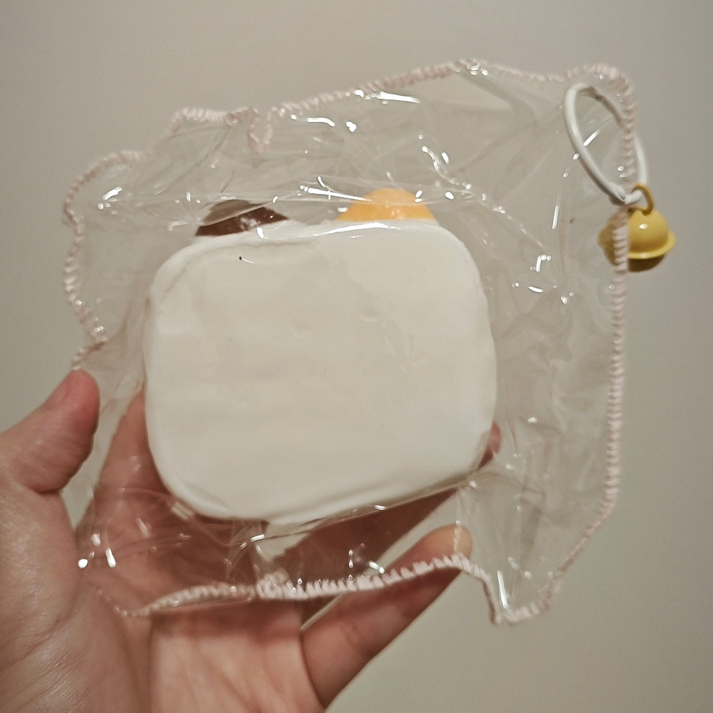 A hand holding a small white squishy toy inside a clear plastic bag with a ruffled edge and a small yellow bell charm, against a plain light beige background.