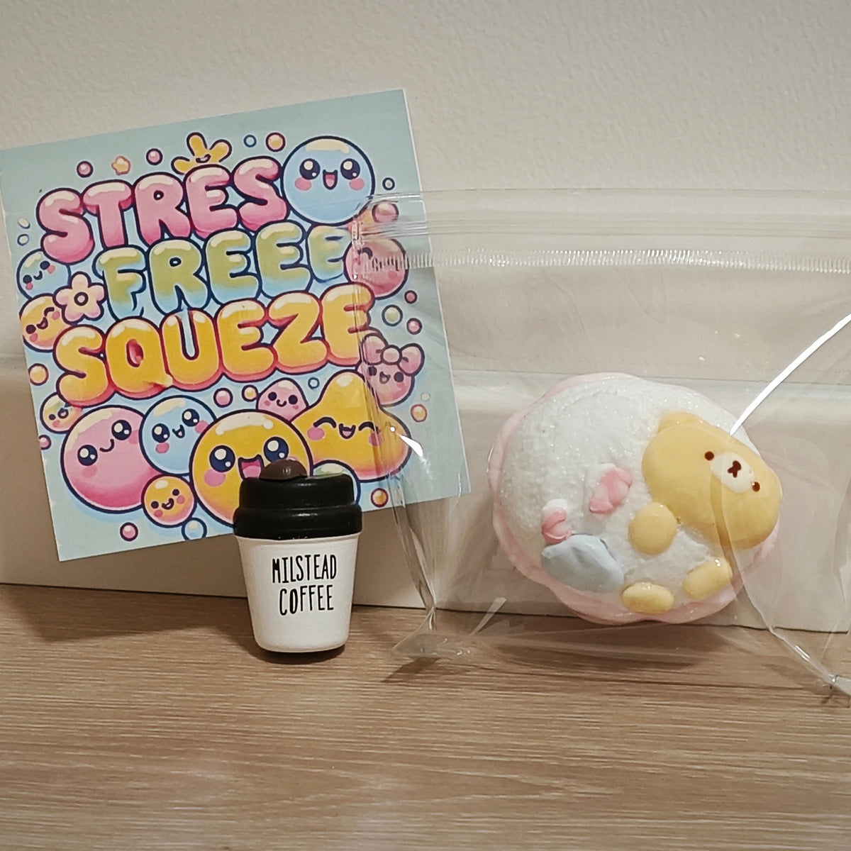 A cute pastel squishy toy with a yellow chick character and pink accents, sealed in a clear plastic bag next to a small coffee cup accessory and a colorful 'Stress Free Squeeze' card on a wooden surface.
