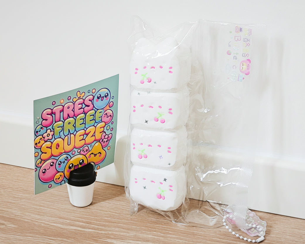 A set of four white squishy toys with pink cherry faces, packaged in a clear plastic bag next to a 'Stress Free Squeeze' card and a small cup, on a wooden floor against a white wall.