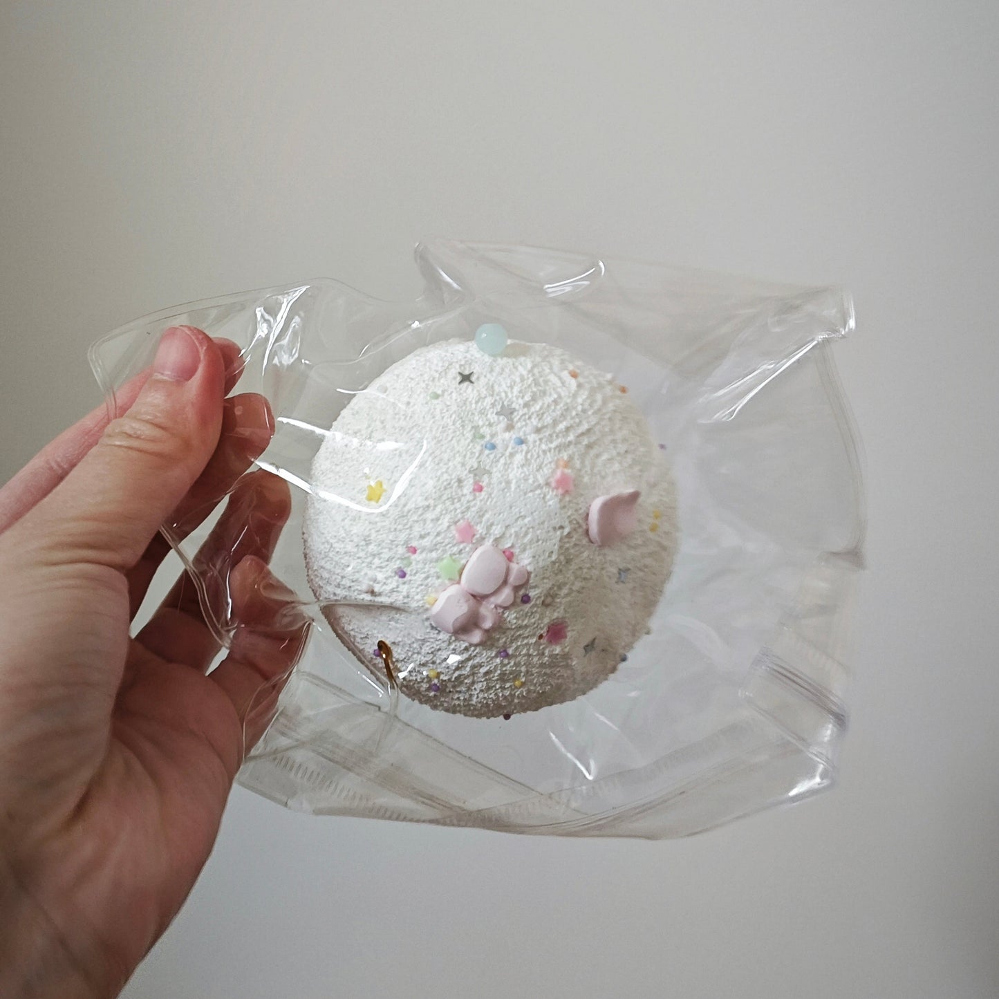 A hand holding a clear plastic bag containing a round, textured white item with small pastel speckles and pink paw-print details, against a plain light gray background.